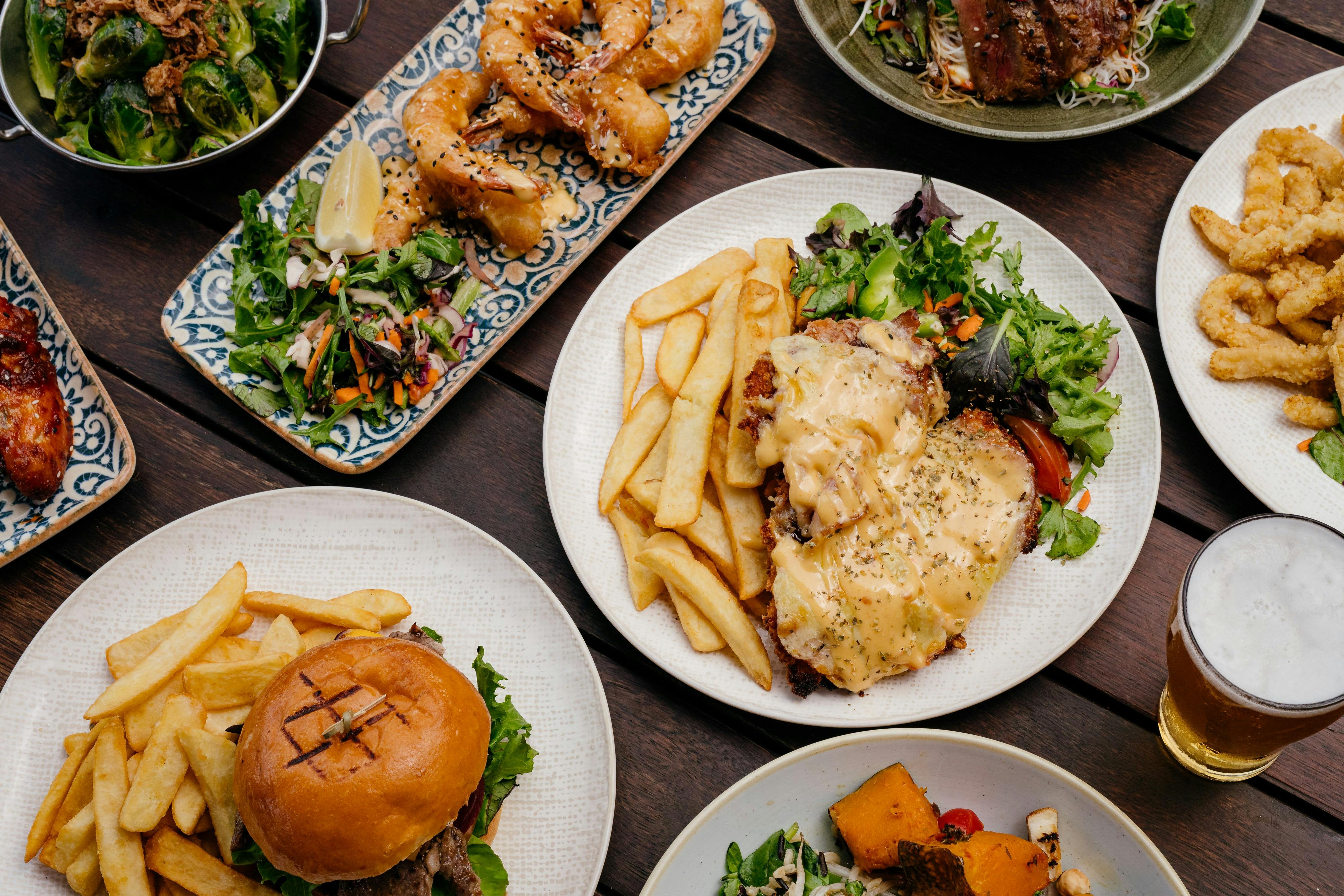 Overhead view of a wooden table spread with a burger, parmi, chips, prawns, and beer.