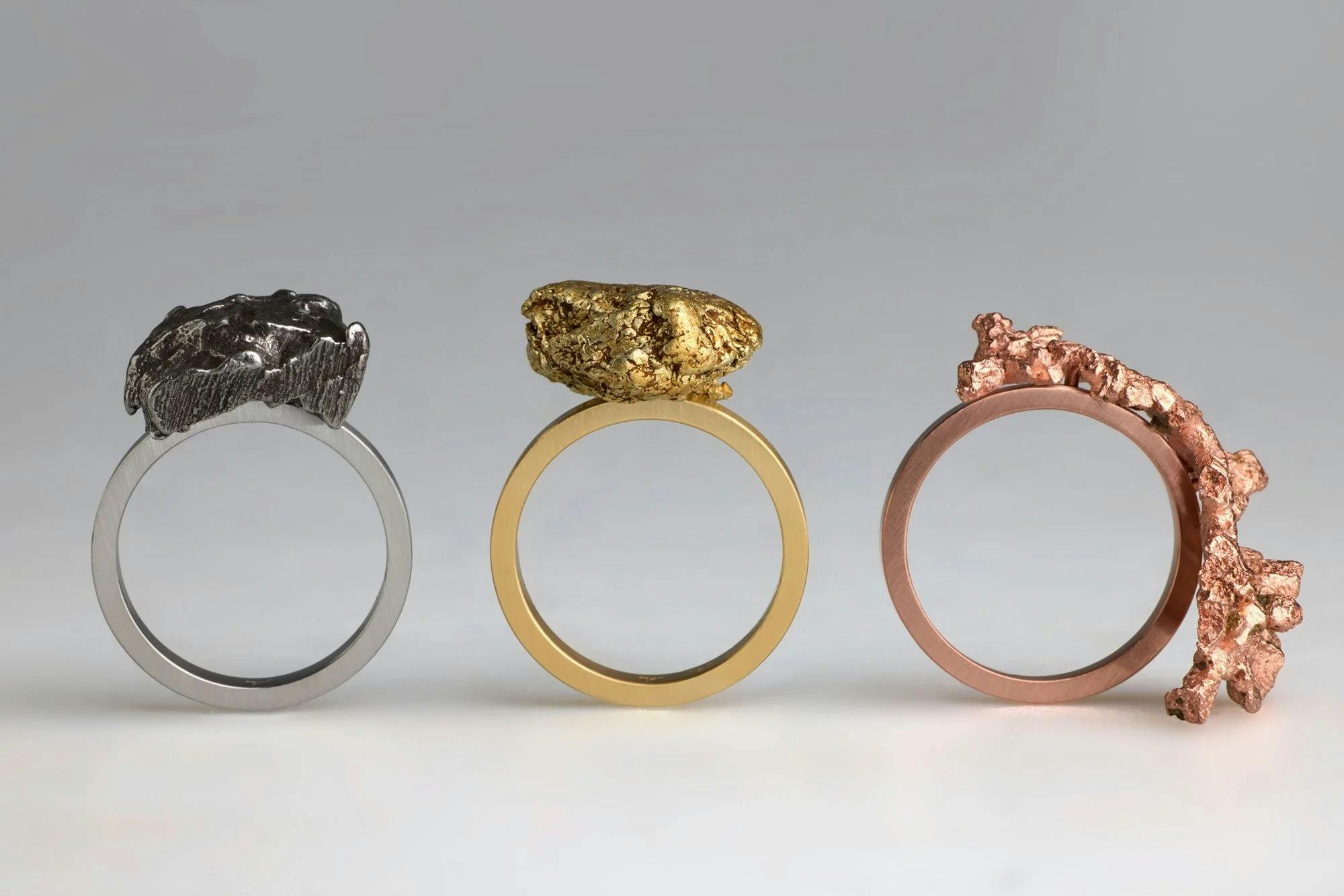 Three rings with raw lumps of metal stand upright; one silver, one gold and one copper