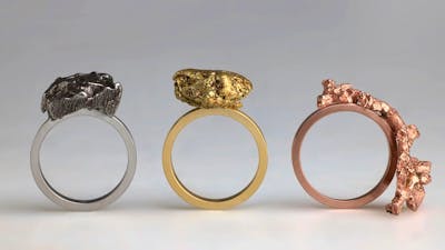 Three rings with raw lumps of metal stand upright; one silver, one gold and one copper