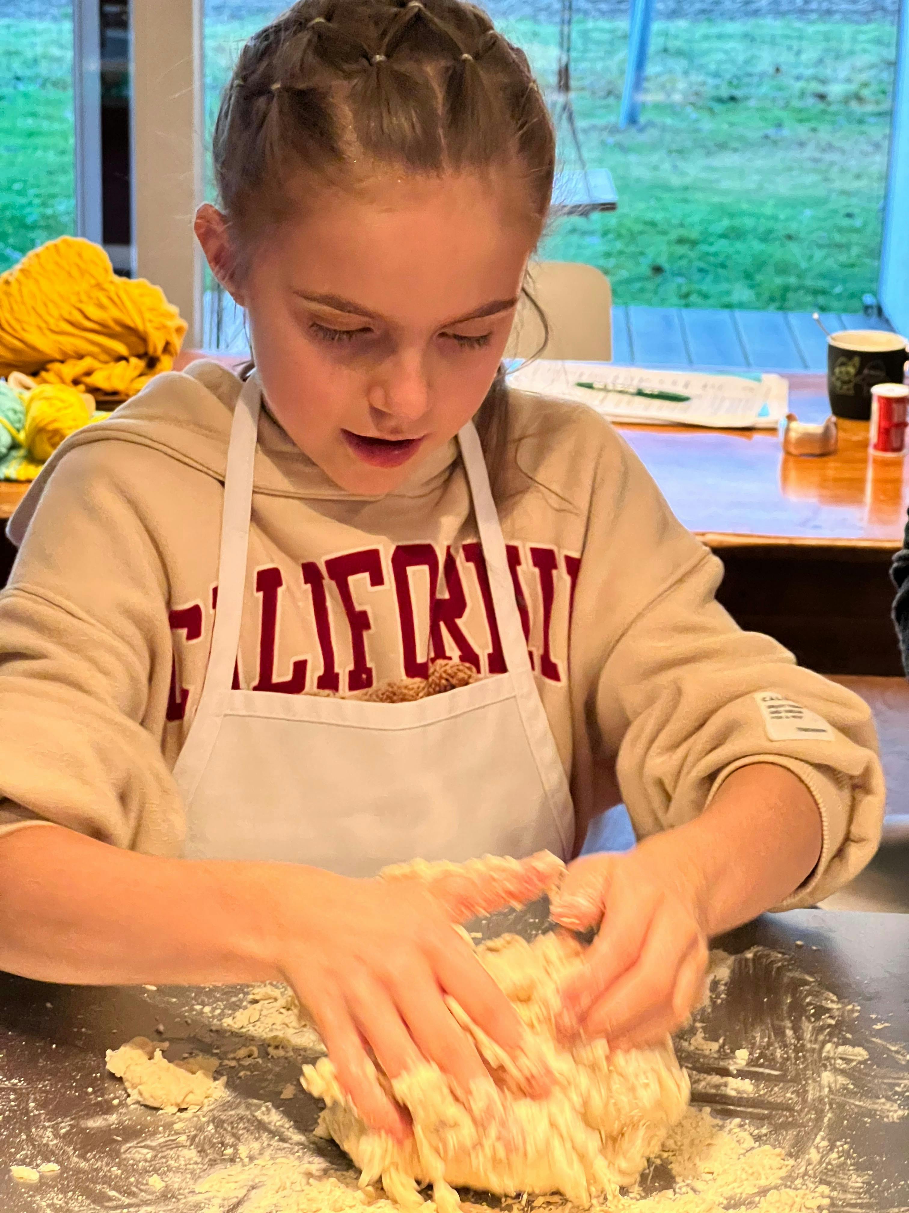 A young participant wearing a white apron works with dough at a kitchen bench during a baking class