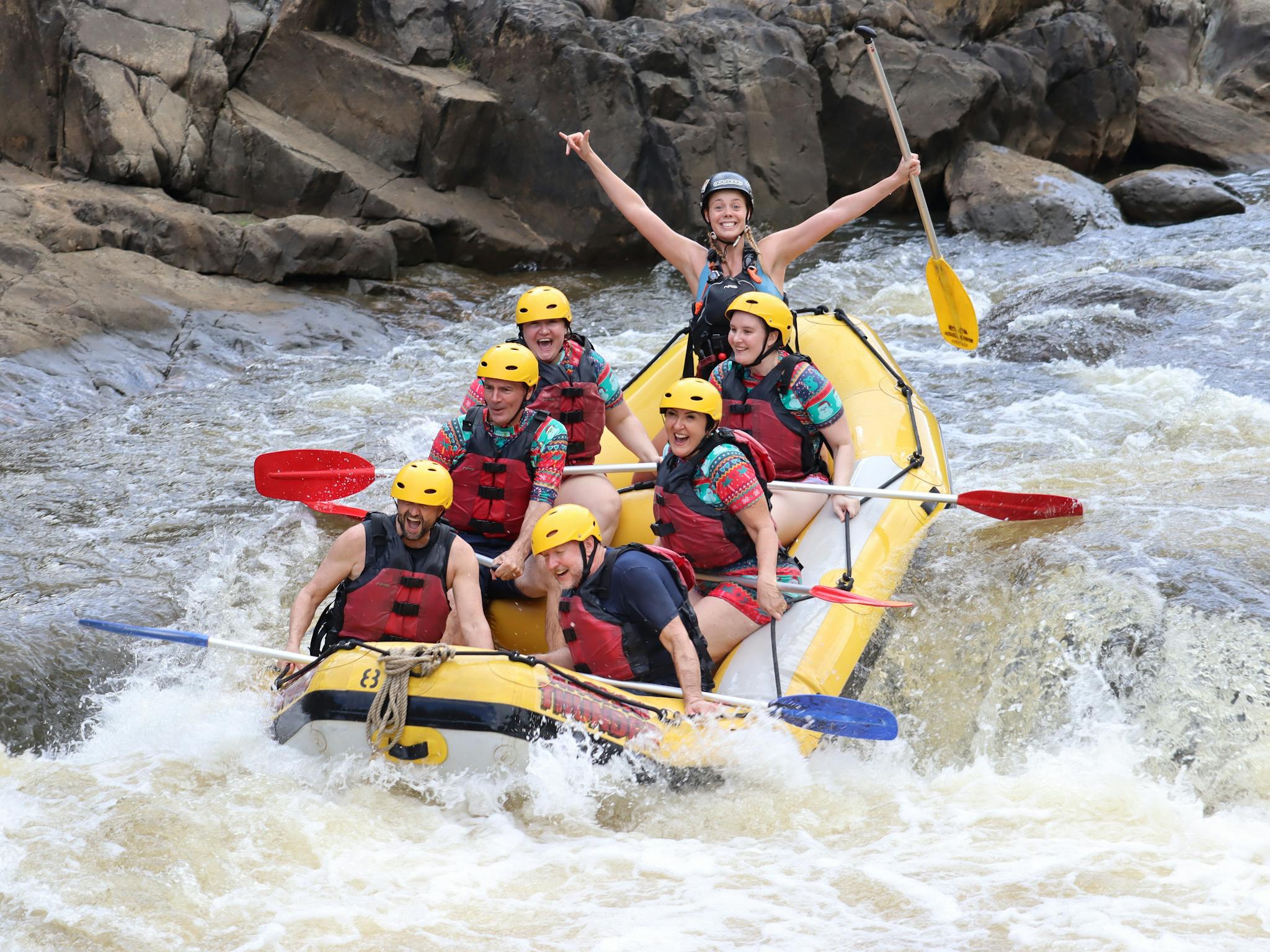 Group of 6 smiling at the camera in the middle of a rapid as their guide lifts both arms victorious Group of 6 smiling at the camera in the middle of a rapid as their guide lifts both arms victorious