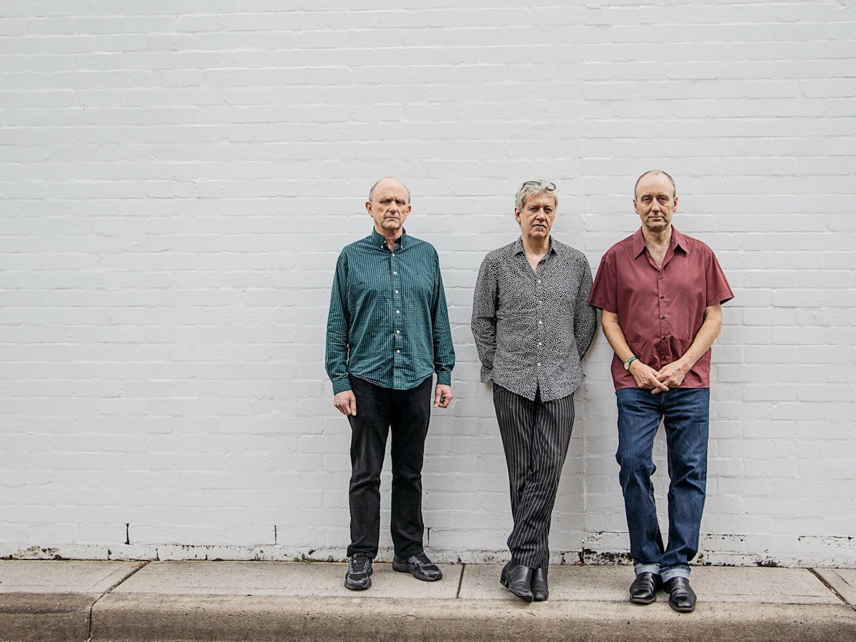 image of three men, standing infront of a grey wall. They are the three members of "The Necks"