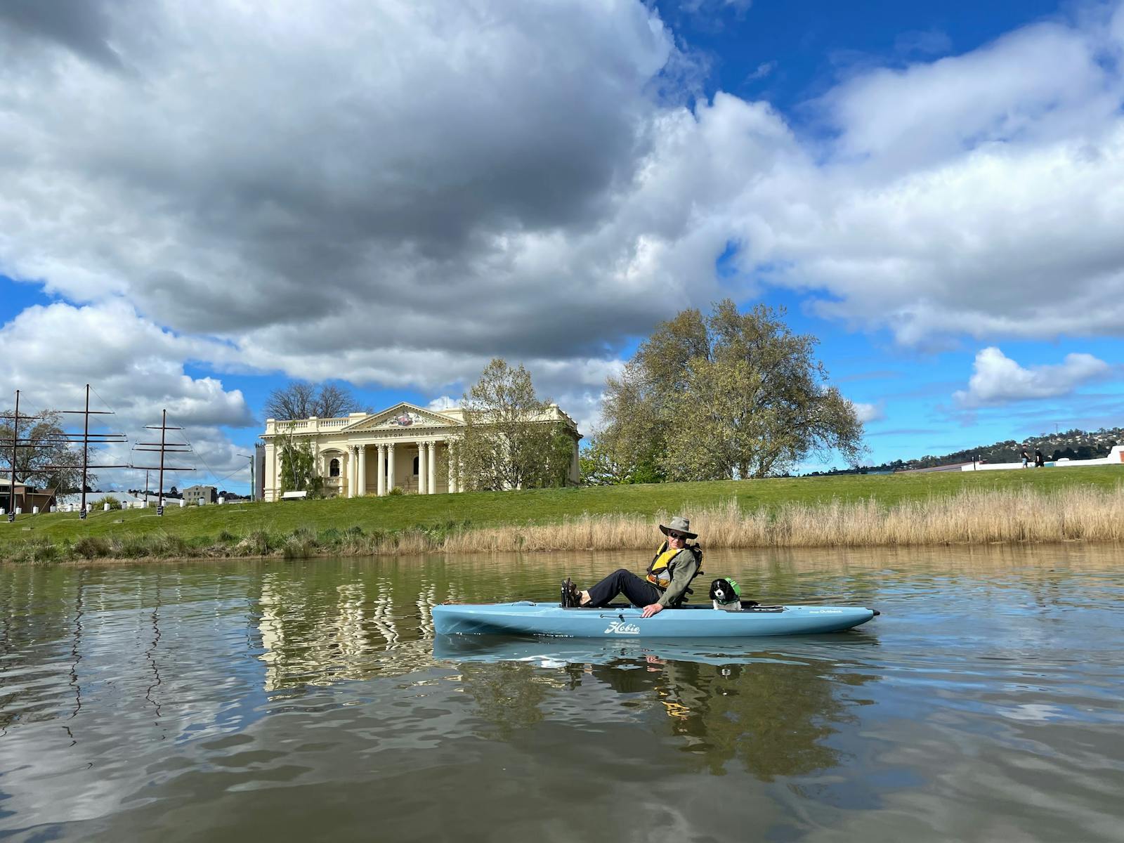A man and a dog in a blue kayak paddle past a old building on the Launceston Waterfront