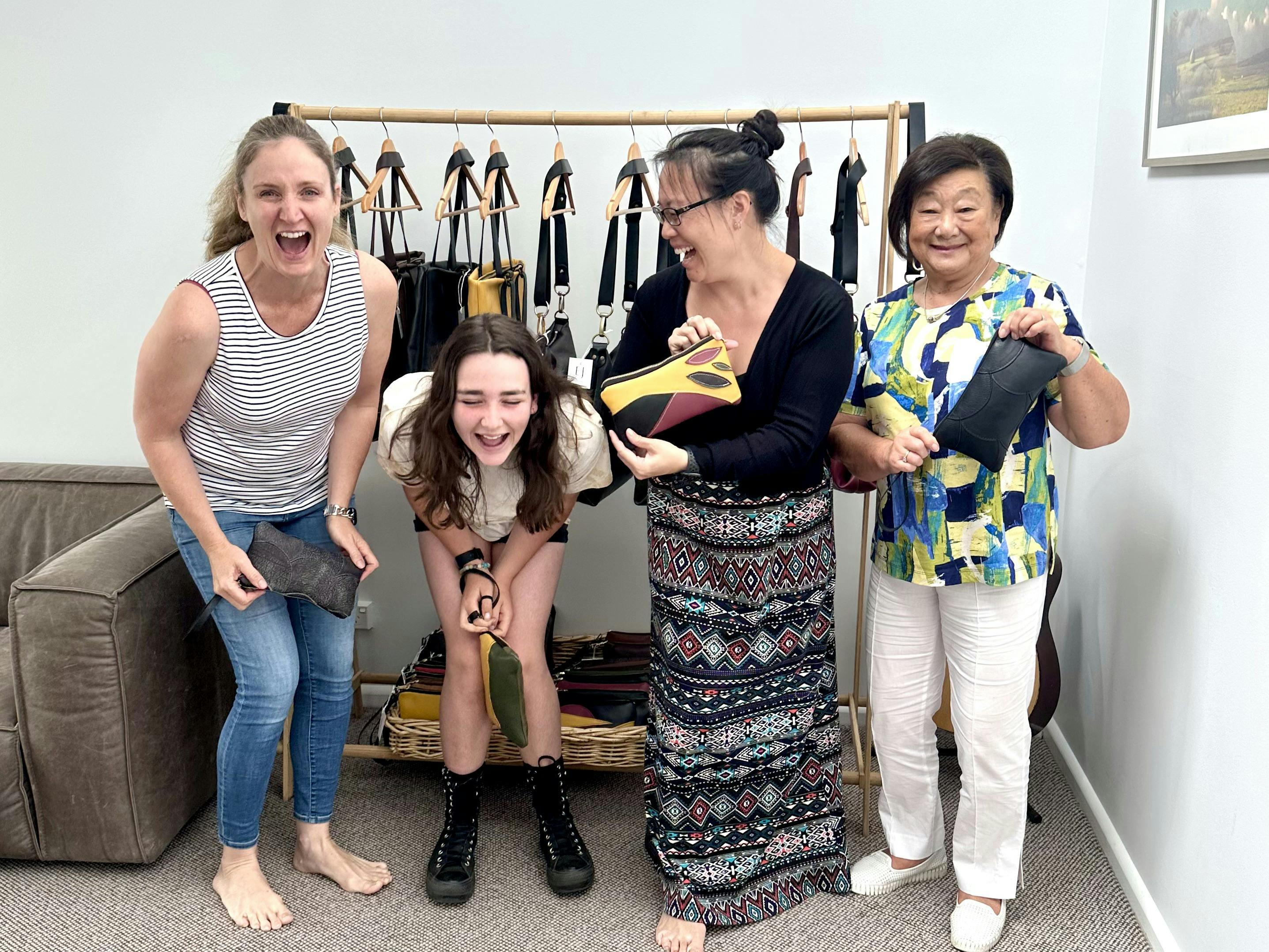 Four women laughing and holding their handmade leather clutches in a workshop setting, with a backdr