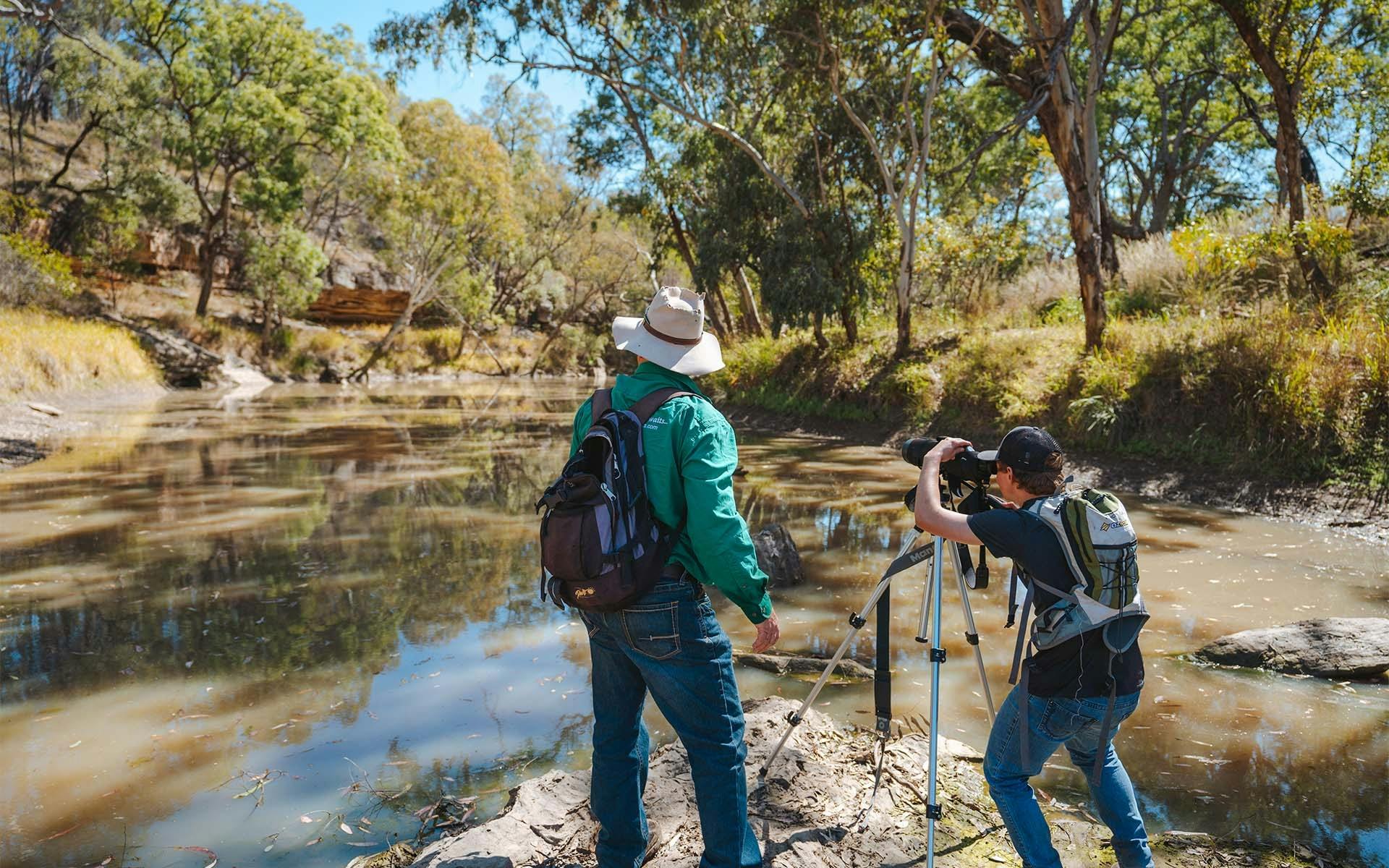 Two men standing on the riverbank with camera and tripod.