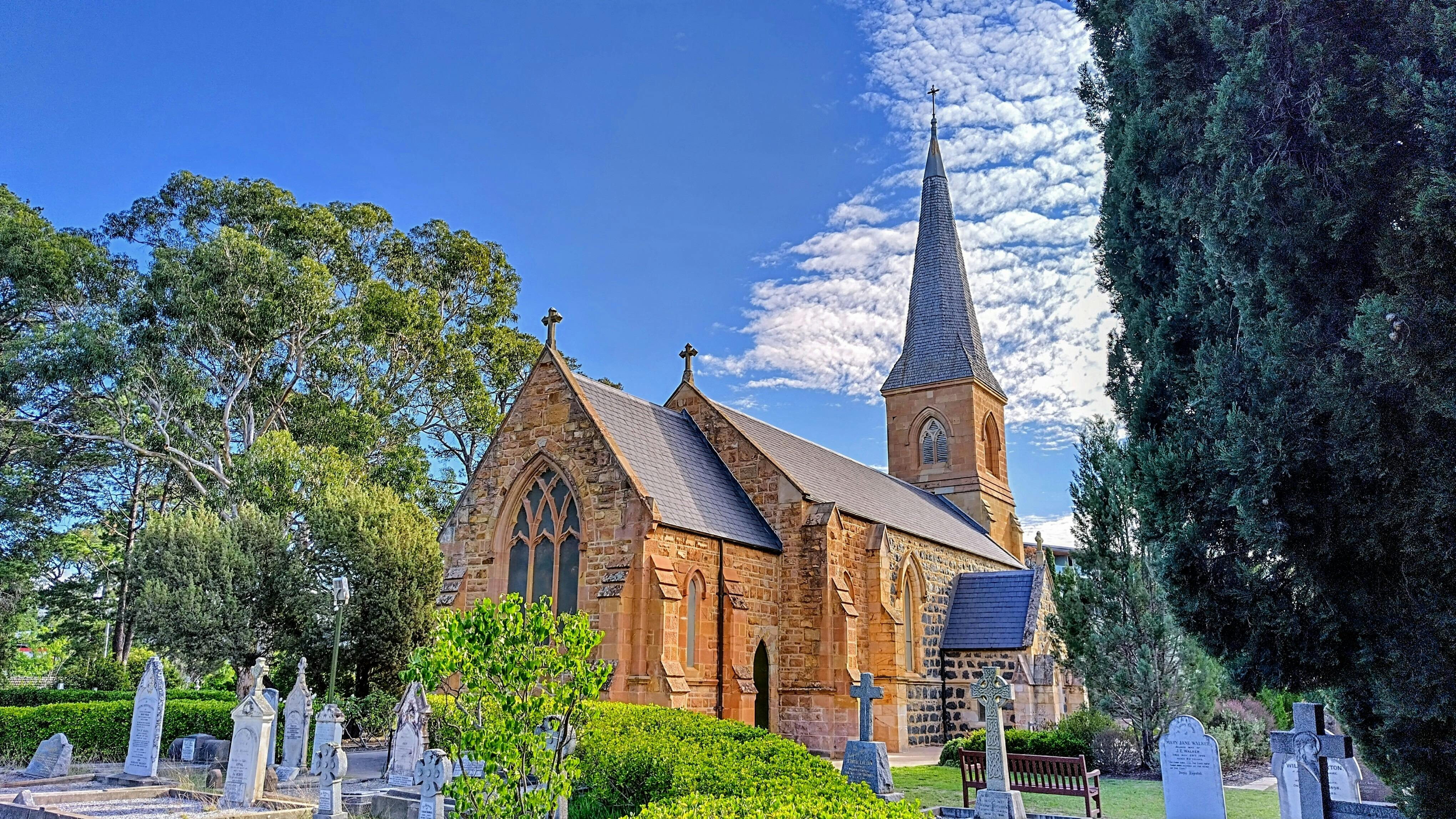 Old graves and gardens surround a sandstone and bluestone church with a spire and bell tower