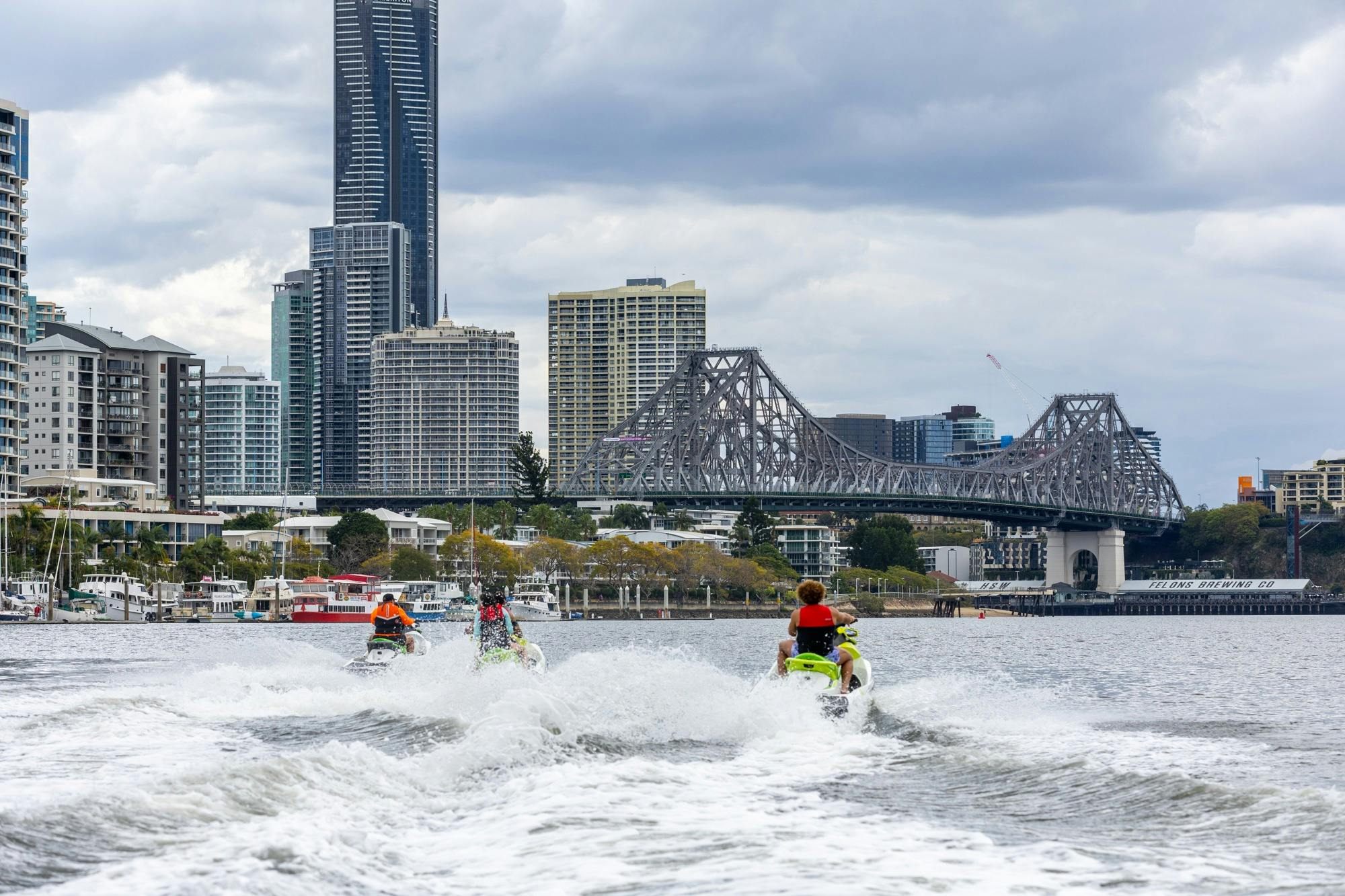 Group of people on jet skis cruising along Brisbane River towards Story Bridge and Brisbane city sky
