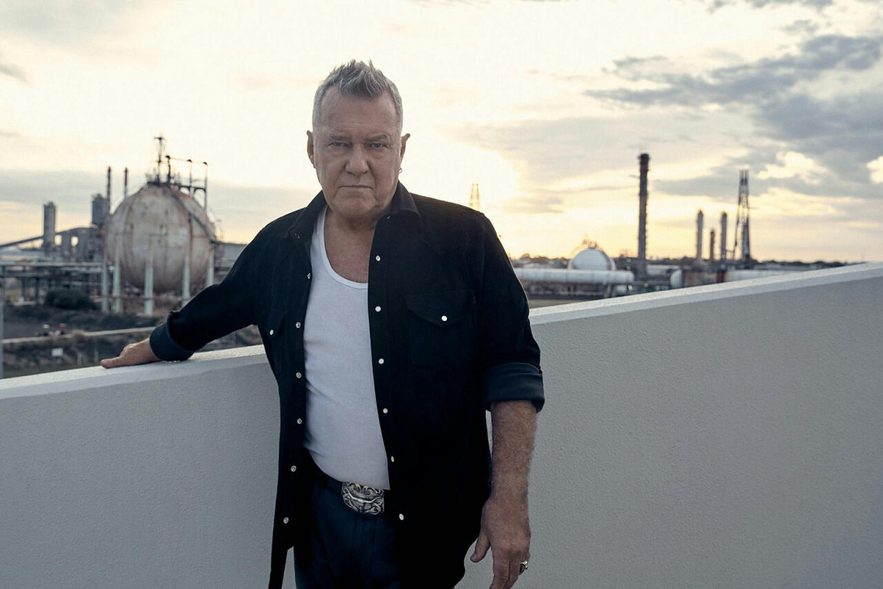 Jimmy Barnes in a black shirt and white singlet, leaning on a wall at sunset.