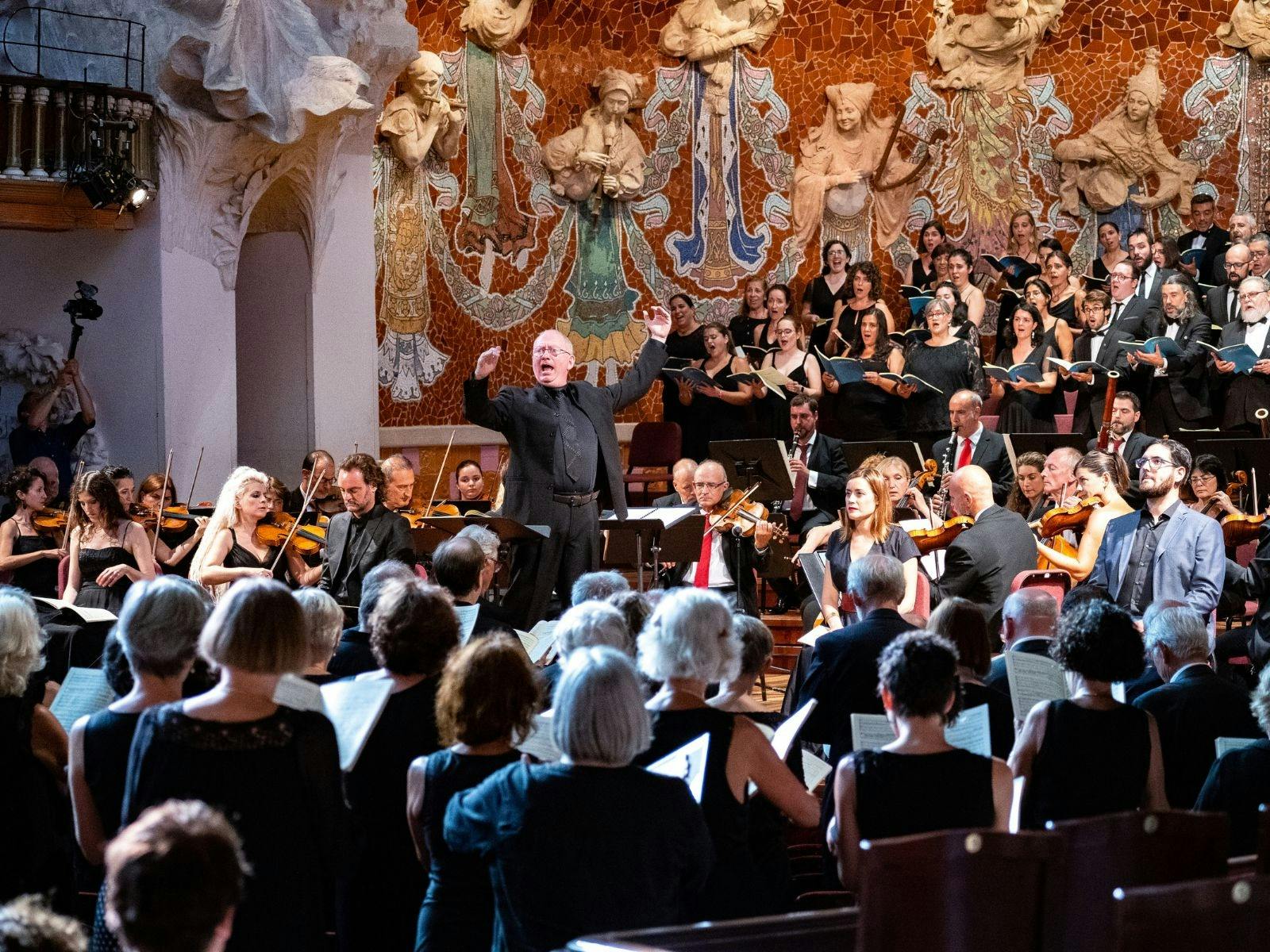 Conductor leading a choir and orchestra in an ornate concert hall with mosaic walls.