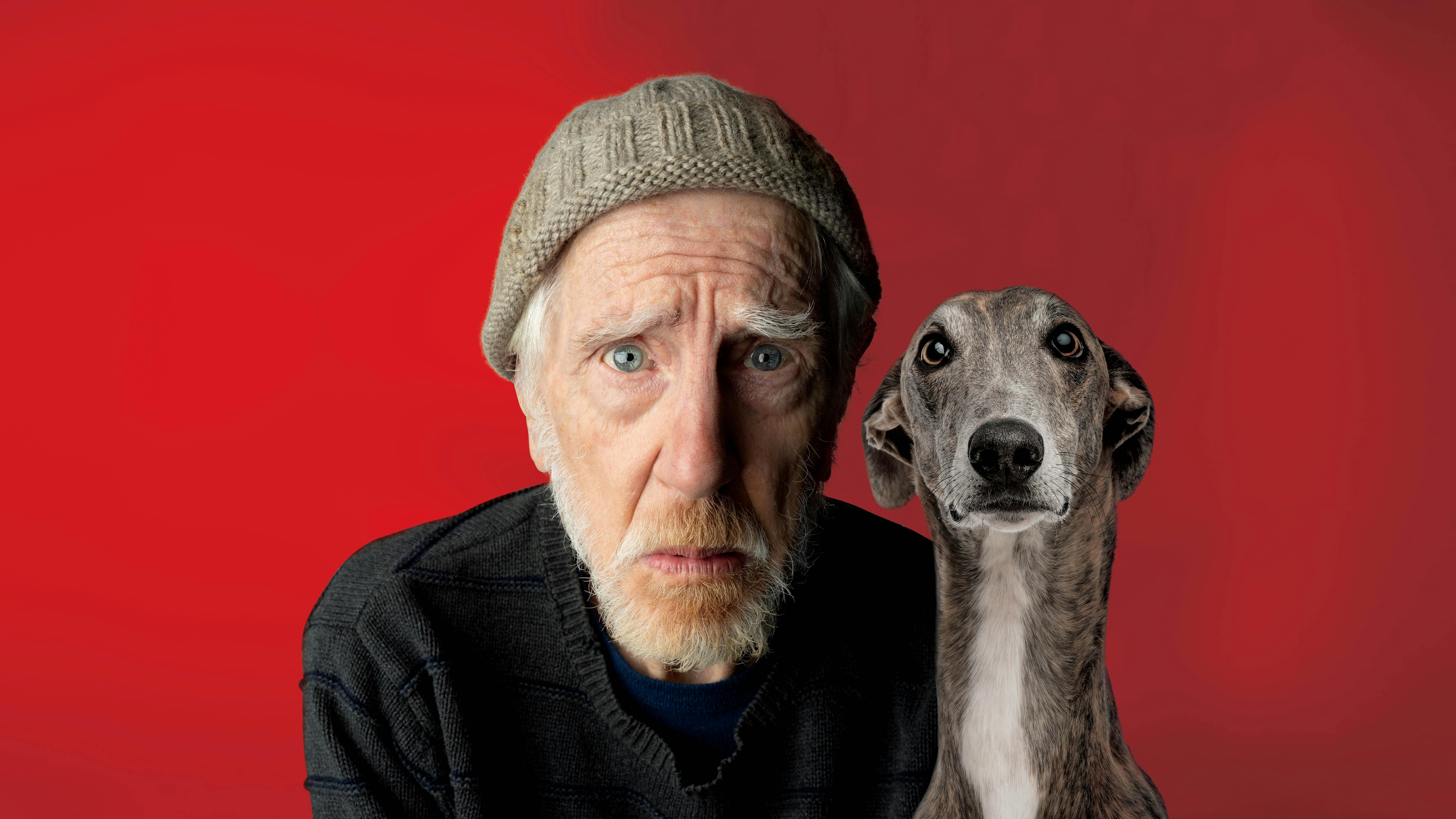 Old man in beanie with a sad face next to a greyhound
