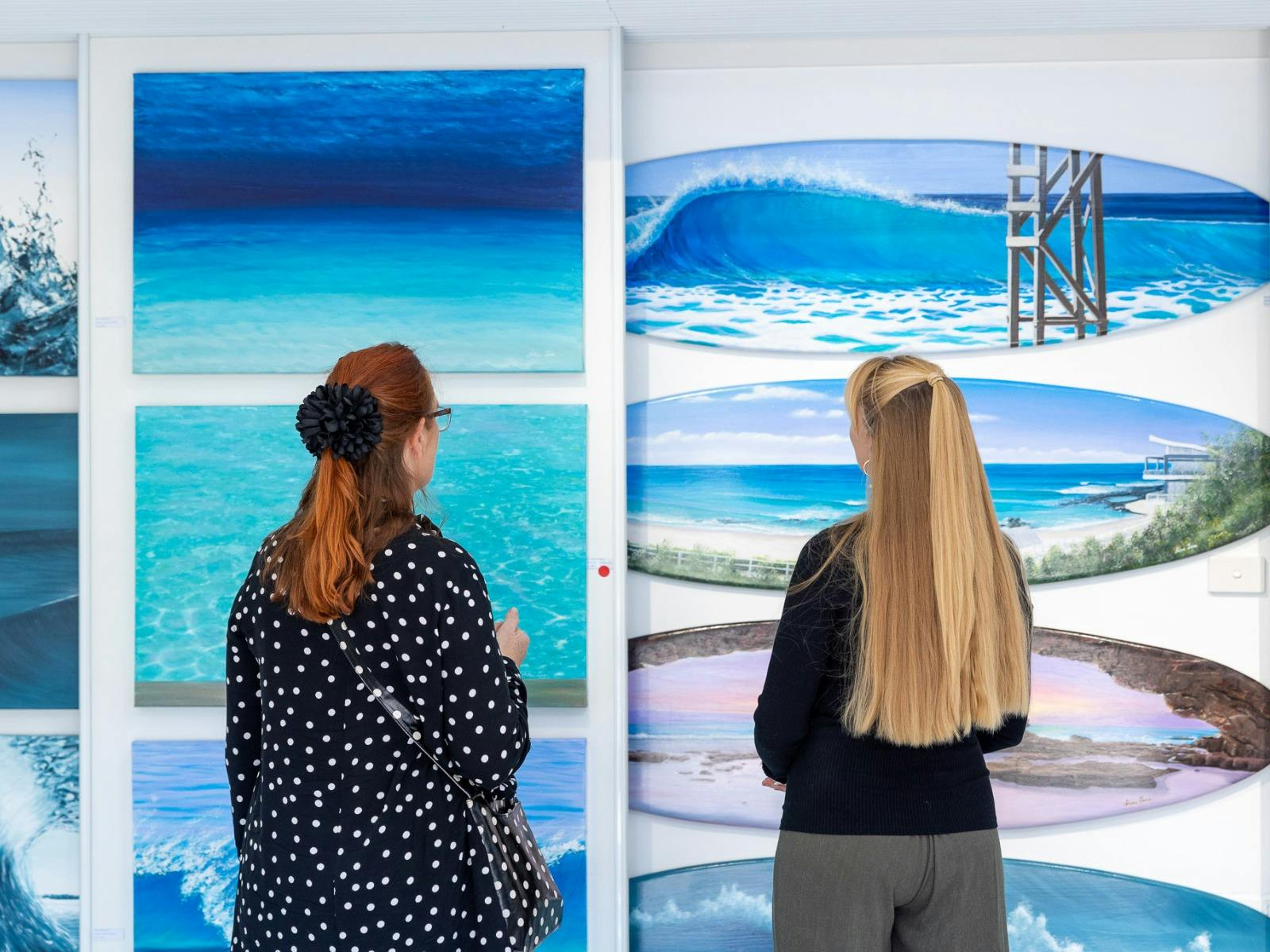 Two people facing away from the camera facing a wall of ocean themed artworks at Lake Mac Open Studi
