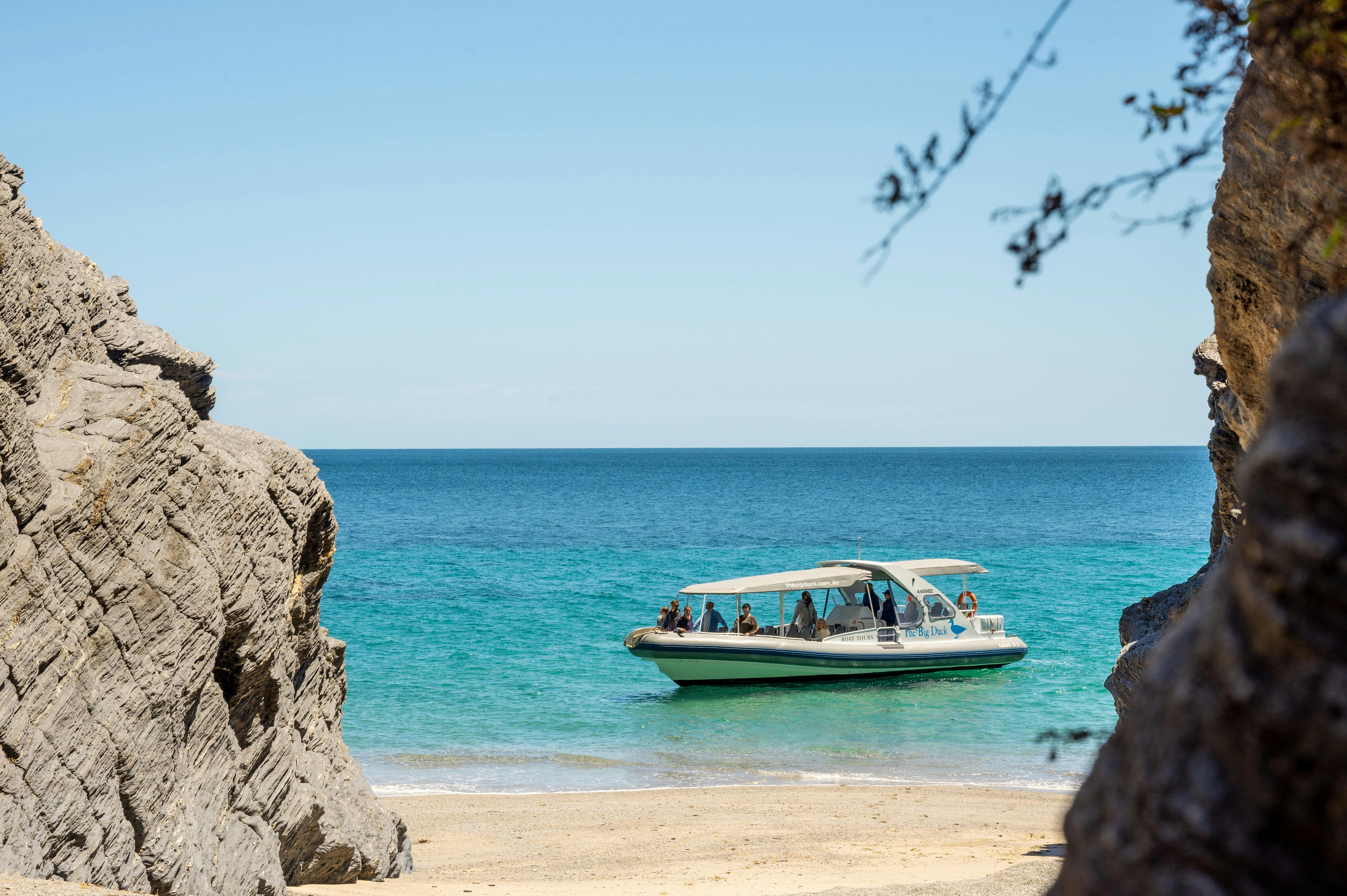 A boat with passengers anchored near a secluded beach, framed by rocky cliffs and clear blue water.