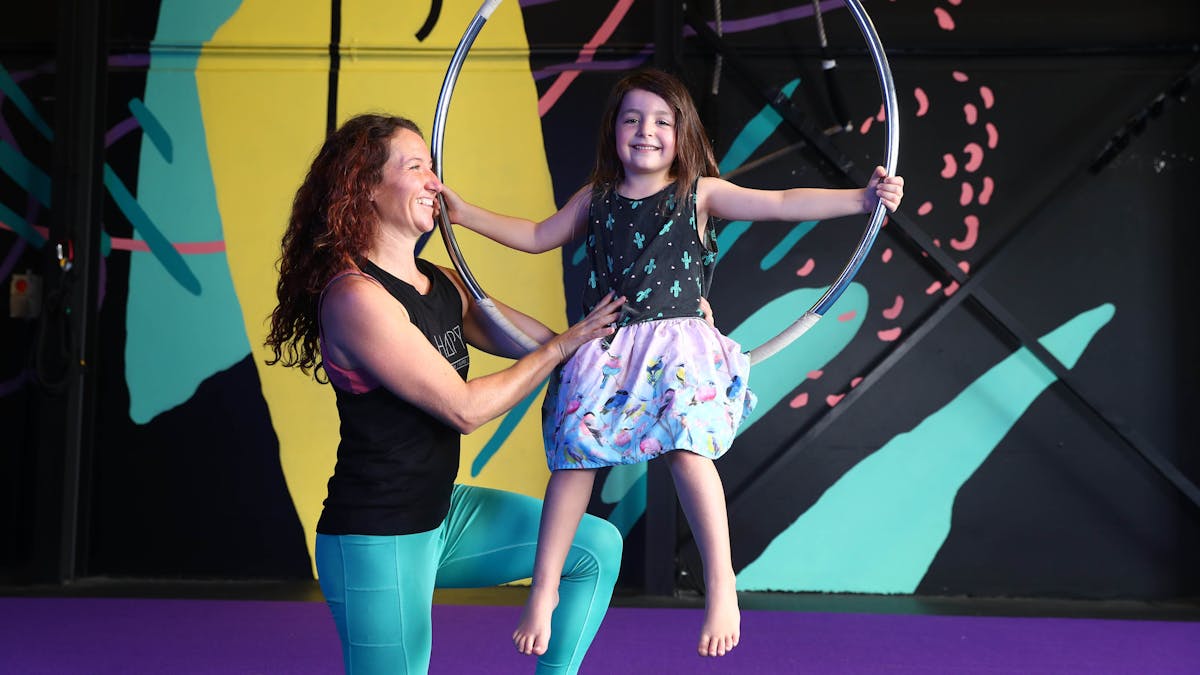 Kids Aerial Classes - Teacher is supporting a  young girl who is sitting on an Aerial hoop.