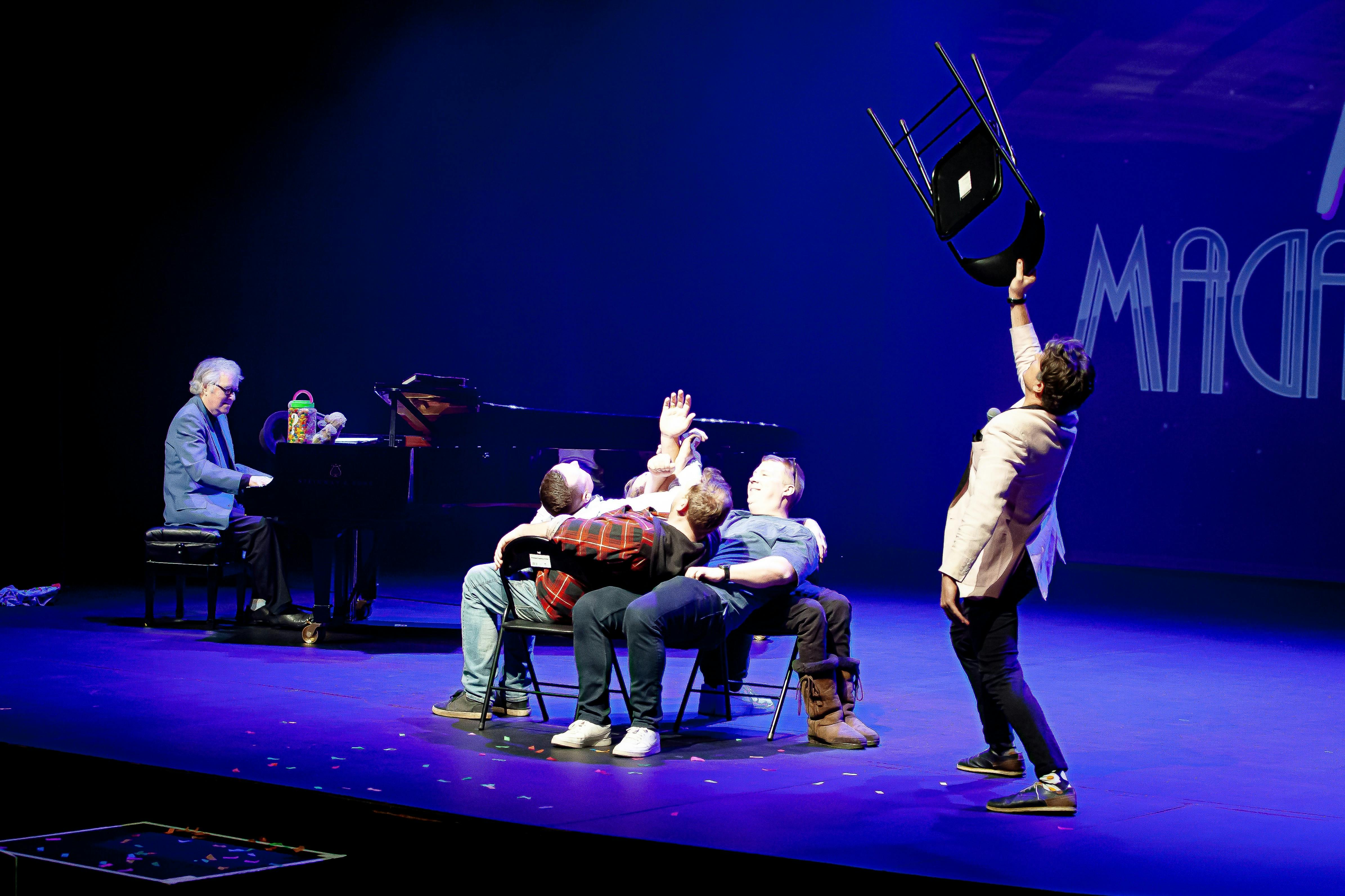 magician adam mada holds up a chair with peter dasent on piano