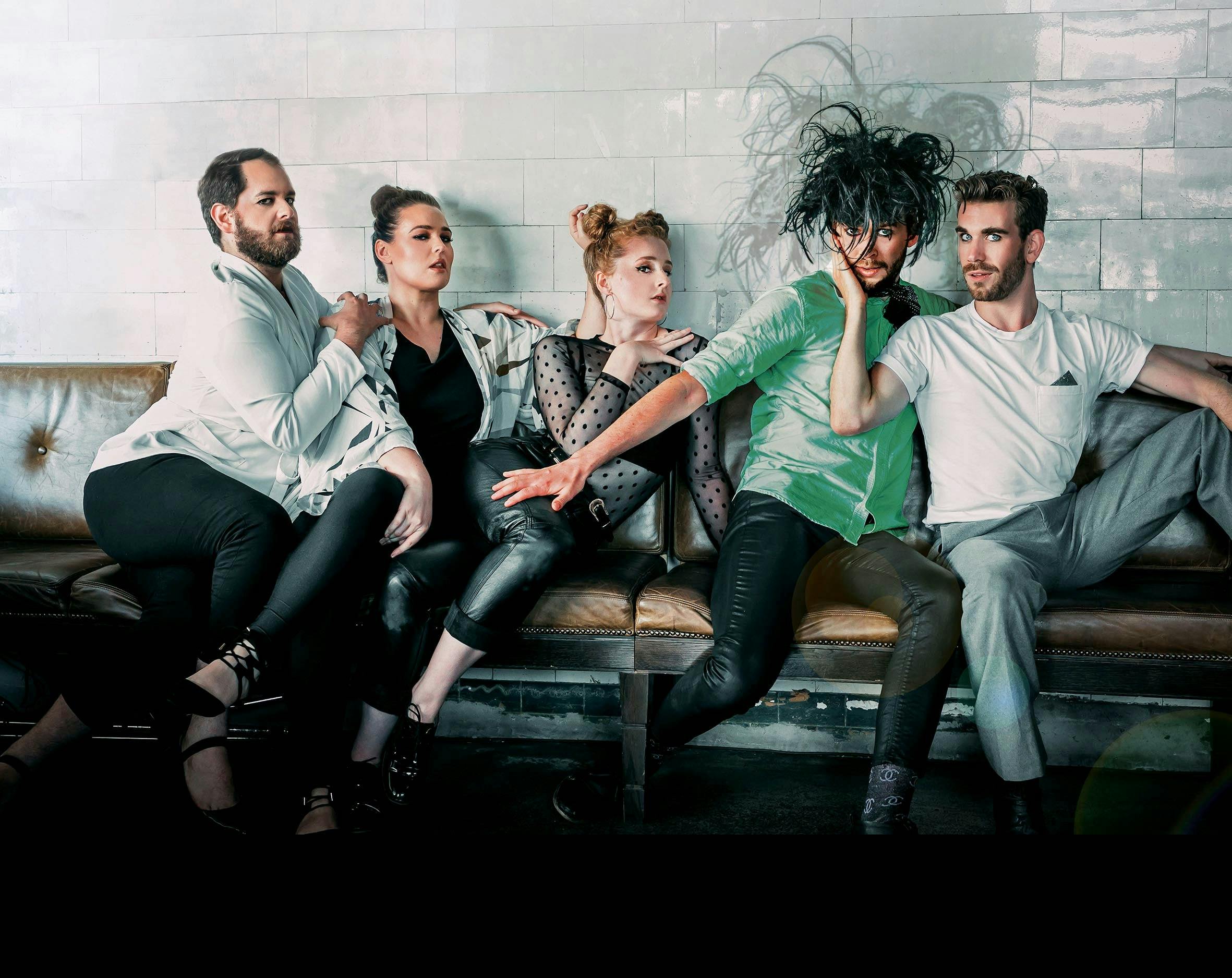 Five people sit on a couch, their hands suggestively on each others shoulders.