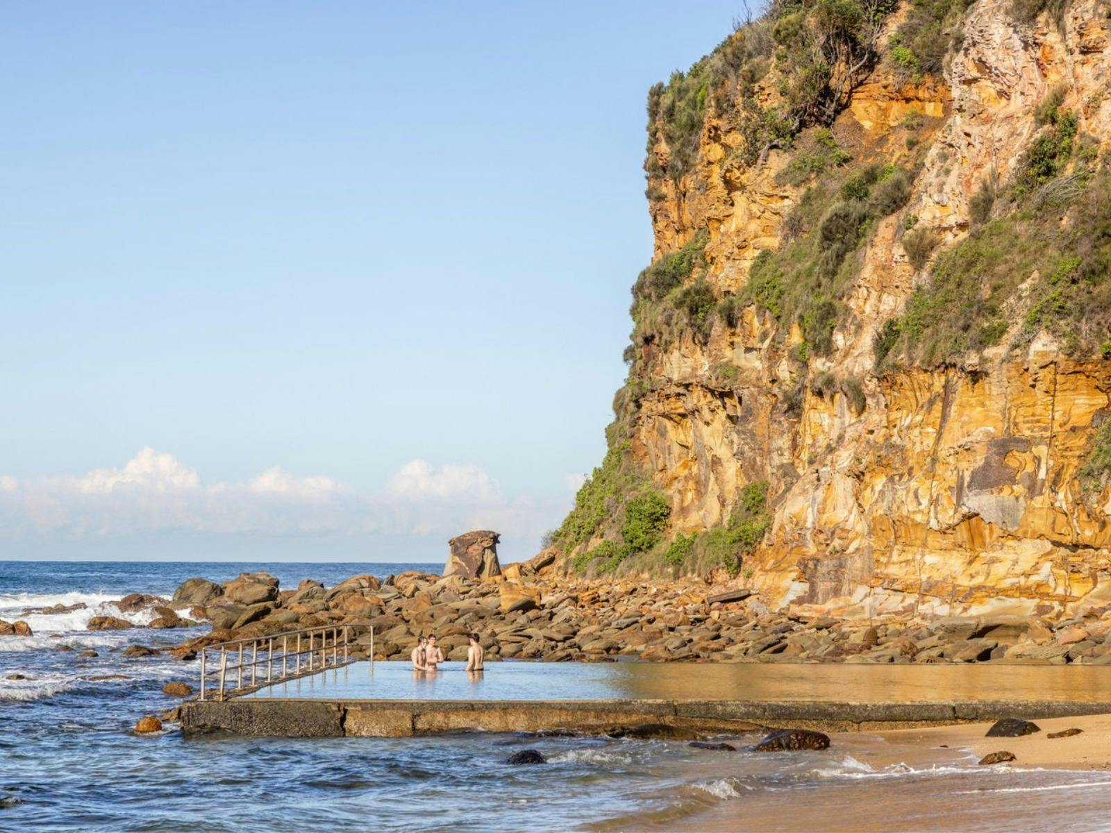 Ocean pool, beneath sandstone cliffs, rocks and ocean, and with people swimming in a pool.