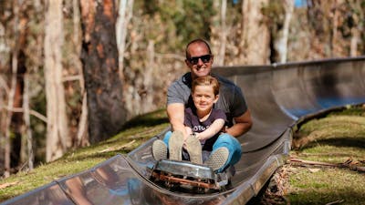 Father and son smiling as they ride on a cart down a metal toboggan track in the forest
