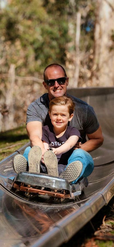 Father and son smiling as they ride on a cart down a metal toboggan track in the forest