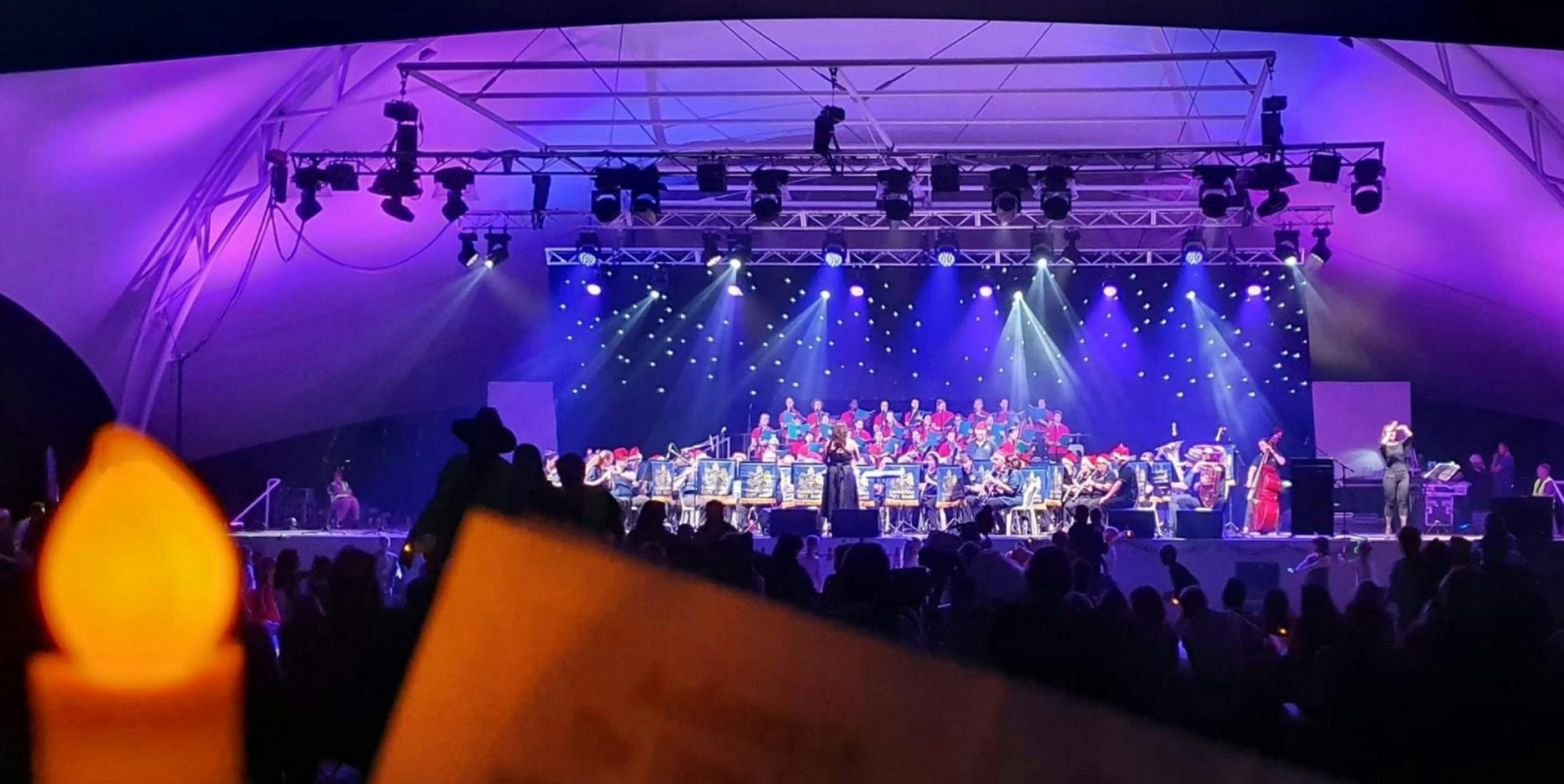 A choir performs on an outdoor stage illuminated by purple stage lights with candles in the audience
