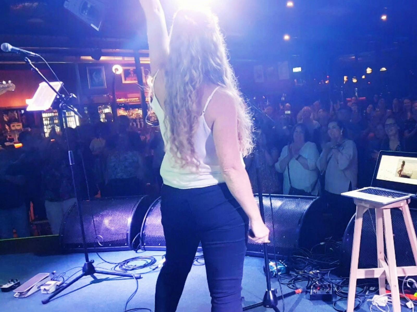 A woman with long blonde hair stands on stage facing a cheering audience, holding a microphone.