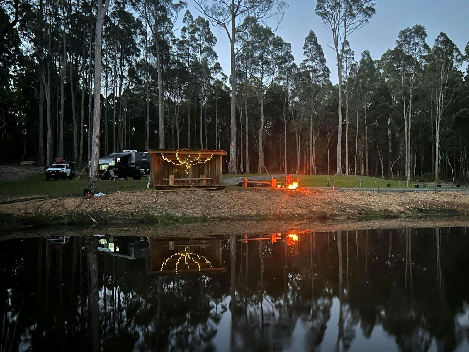 A wooden shelter with fairy lights, a campfire, and tall trees reflecting on a calm water surface at