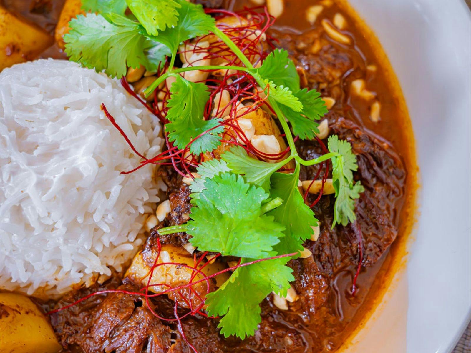 Close-up of beef curry with rice, cilantro, peanuts, and chili threads in a white bowl.