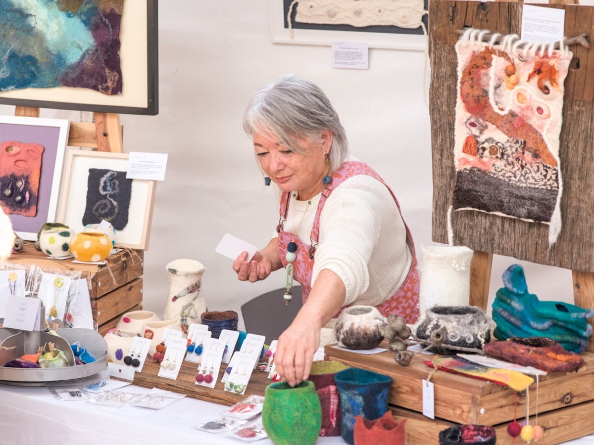 Women in white shirt and pink overalls standing behind stall with artisan earrings and prints