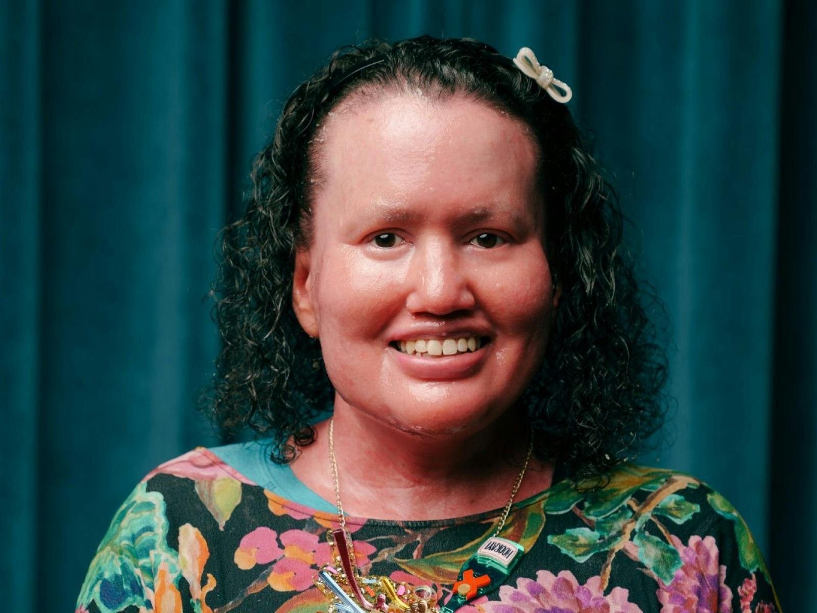 Portrait of Carly Findlay, smiling and wearing a colourful floral top,  in front of a teal curtain