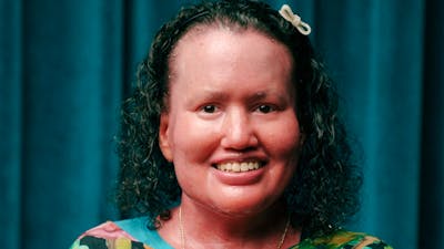 Portrait of Carly Findlay, smiling and wearing a colourful floral top, in front of a teal curtain
