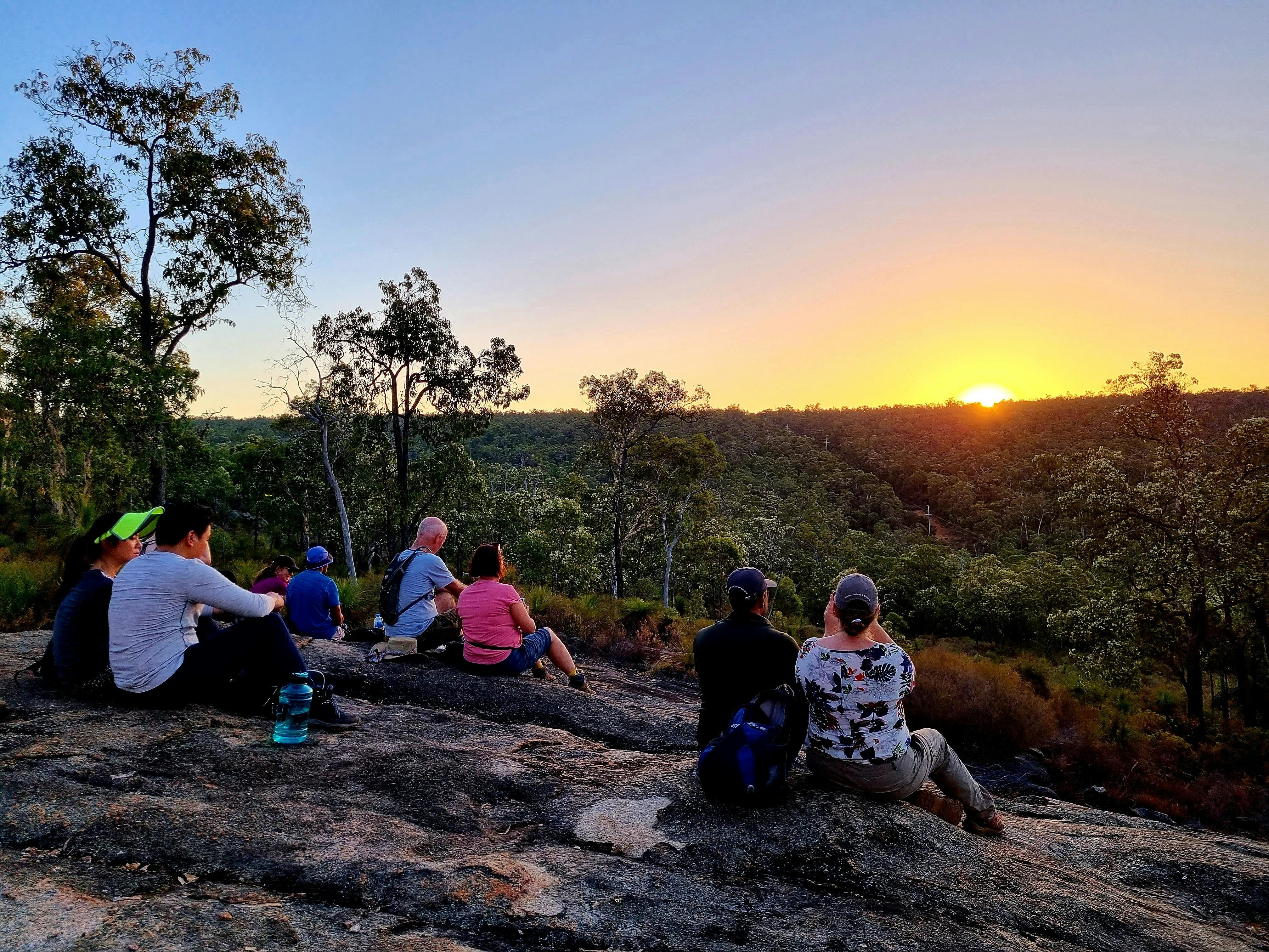 Hikers sitting on top of hill as sun sets below hills in background, warm yellow and purple hues