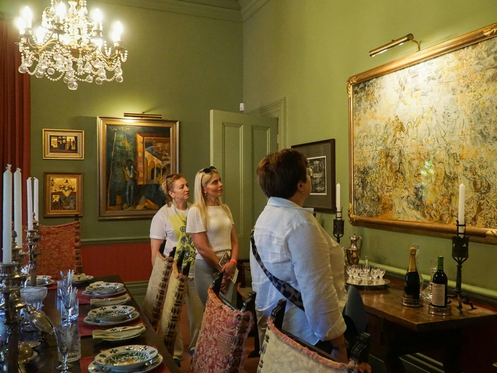 Three women look curiously at a painting. A chandelier is lit and dining table set to their right.