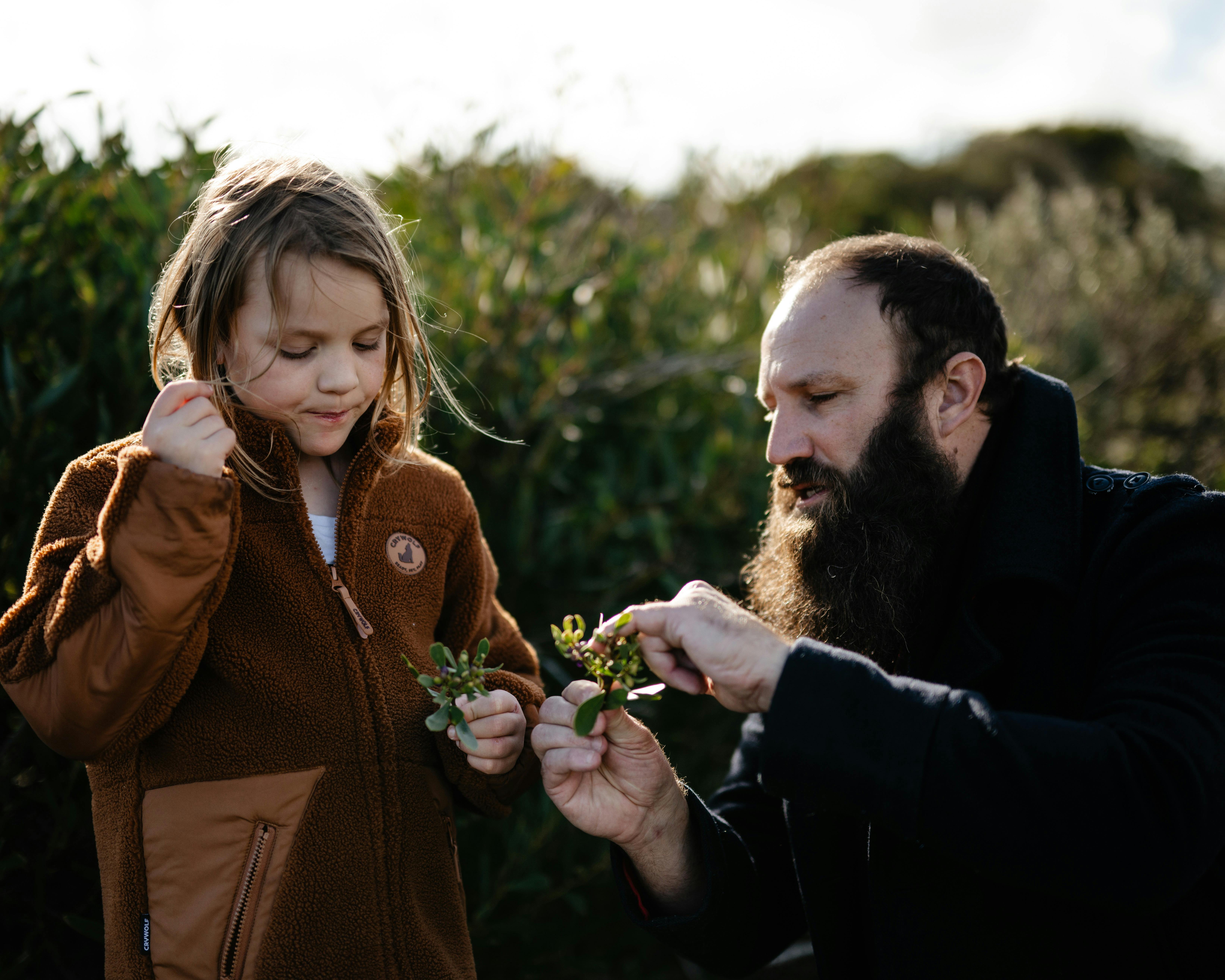 A bearded man shows a plant to a young girl in a brown jacket, both exploring nature together.