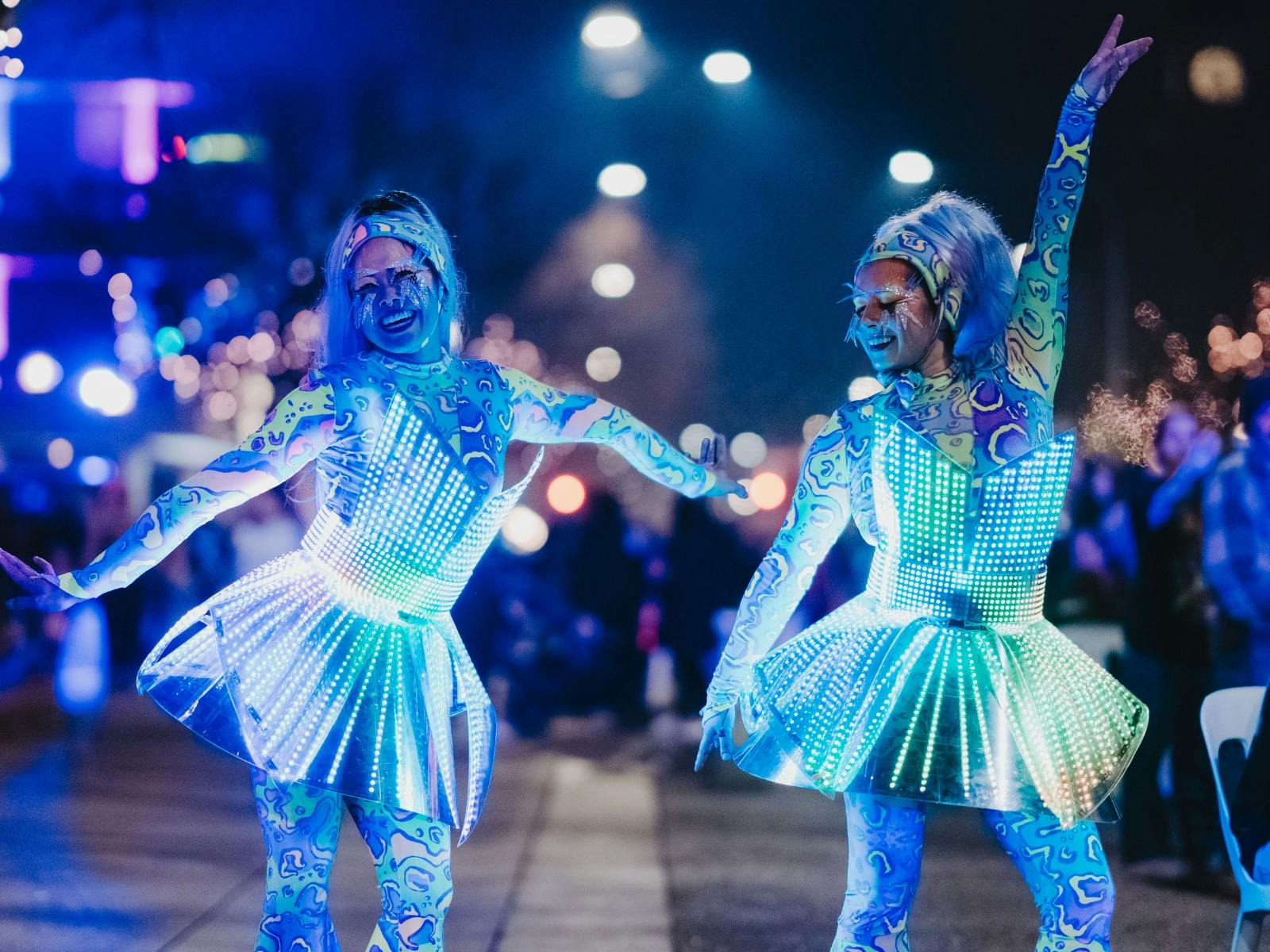 Two performers in bright LED outfits dancing under blue lights at night.