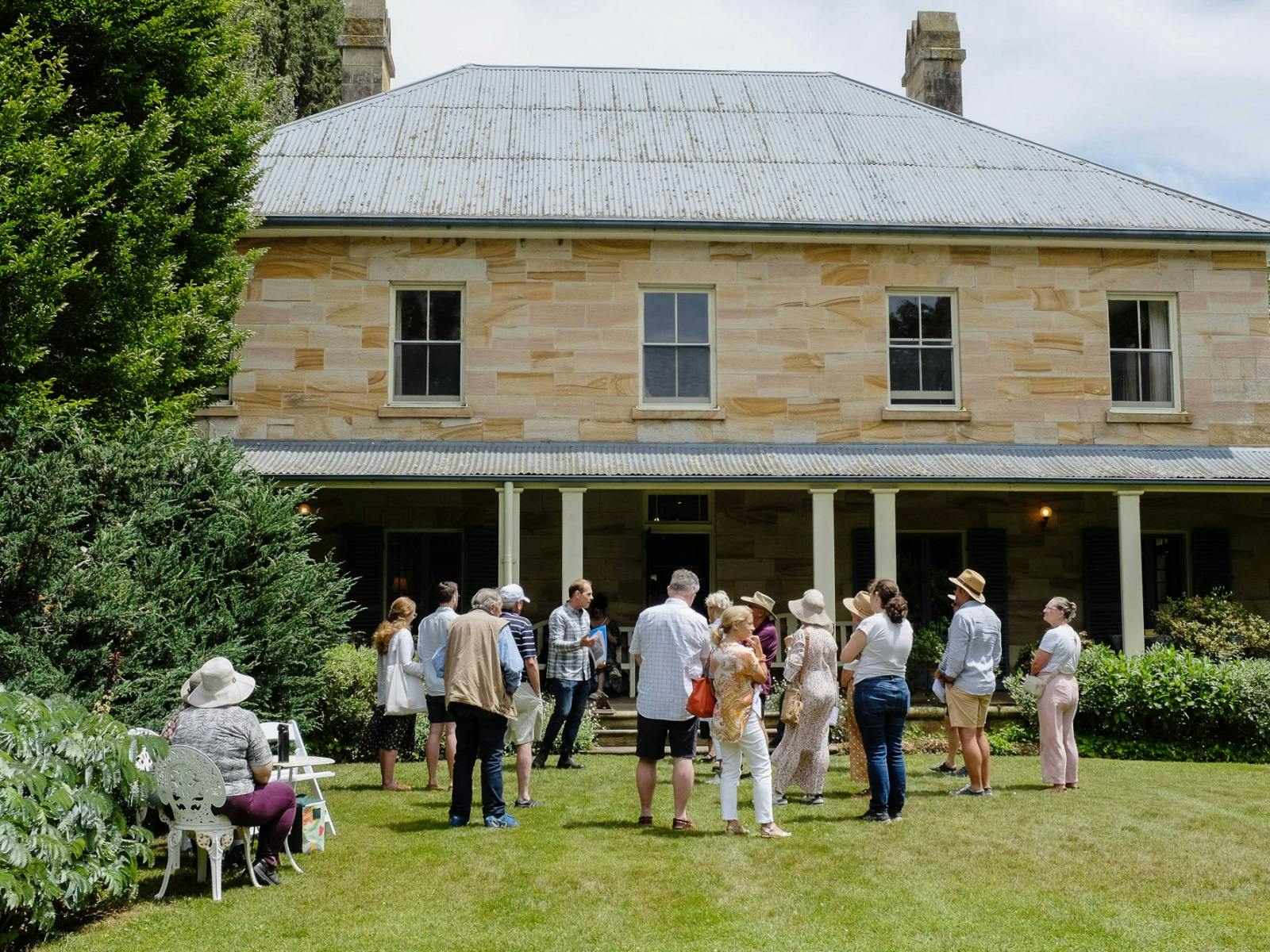 A group of people gather on a green lawn infront of a sandstone building.