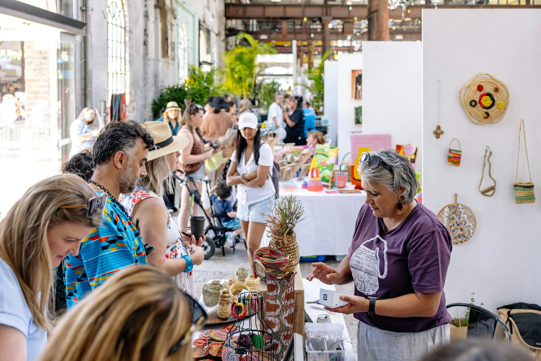 A bustling outdoor market scene with shoppers browsing various displayed items and colourful stalls.