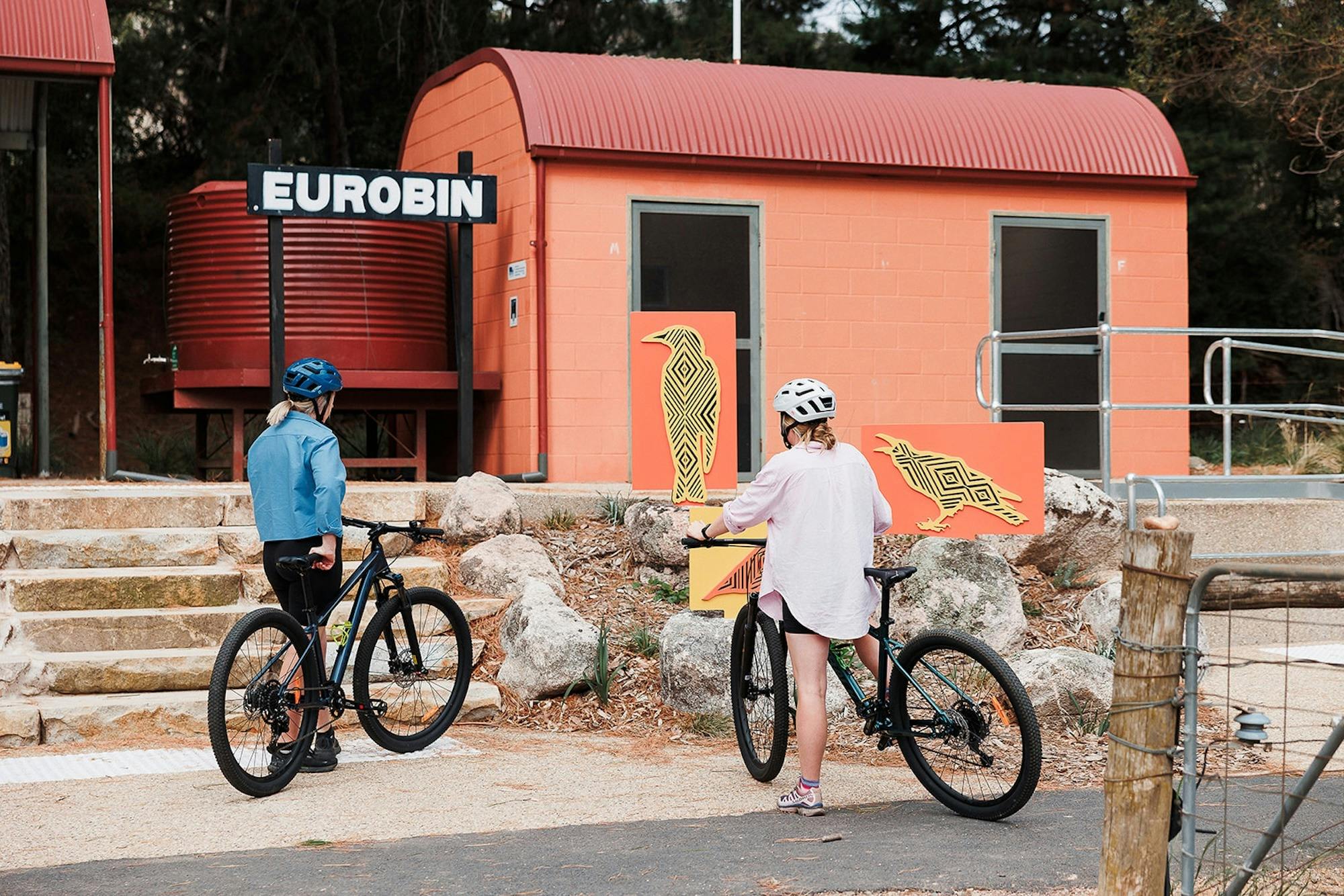 Two cyclists looking at Indigenous art of birds in front of an orange building and 'Eurobin' sign
