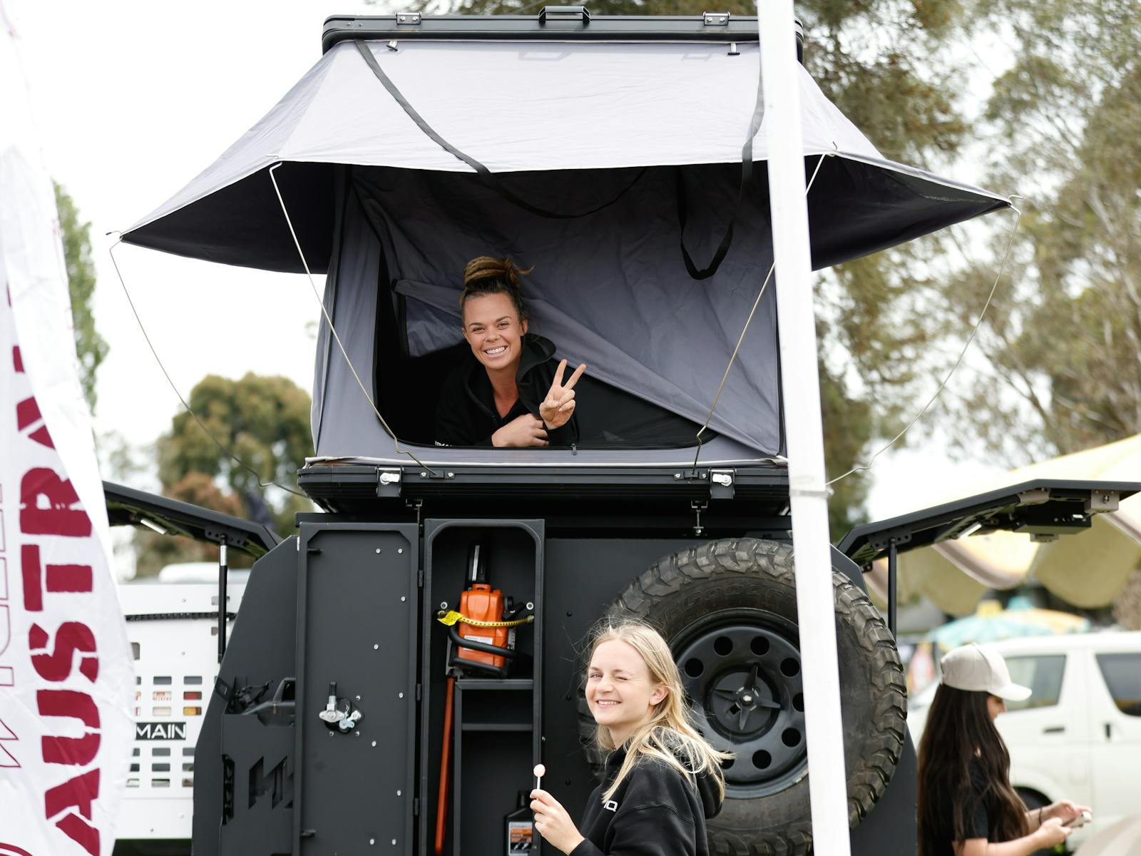 One young women lying in rooftop tent giving peace sign and a second down below with a lollipop