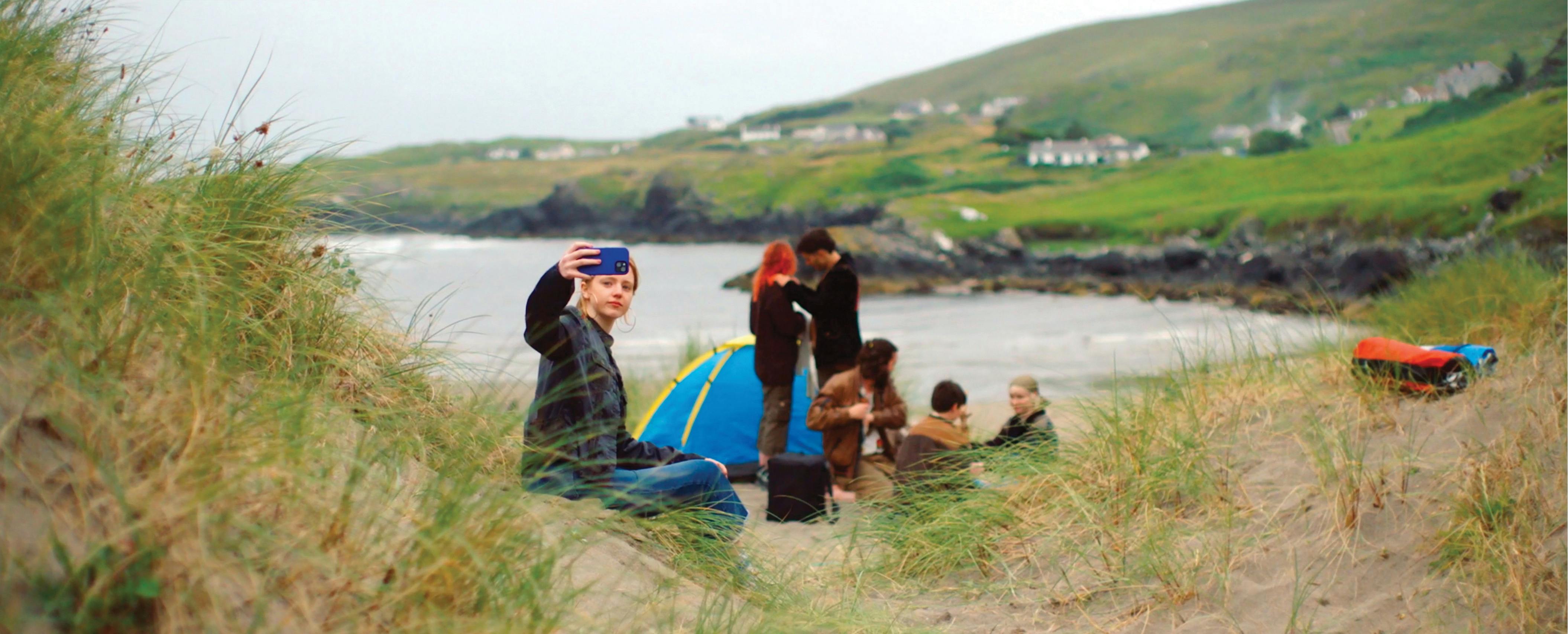 Boy holds up a phone with his family behind him setting up a tent in the Irish countryside