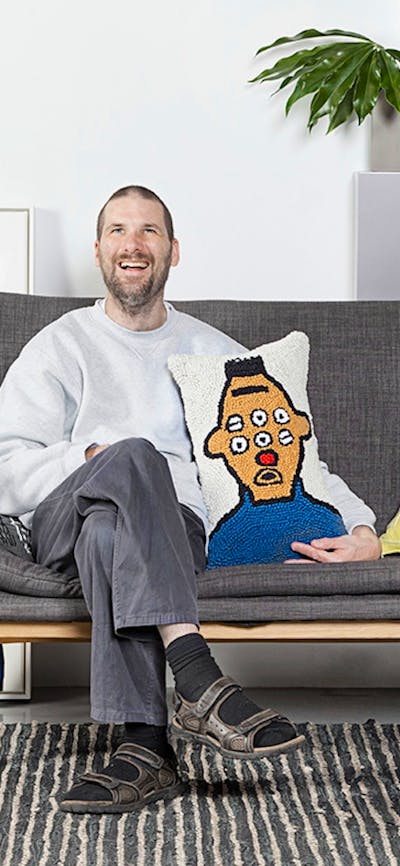 A man sitting on grey couch holding a colourful cushion.