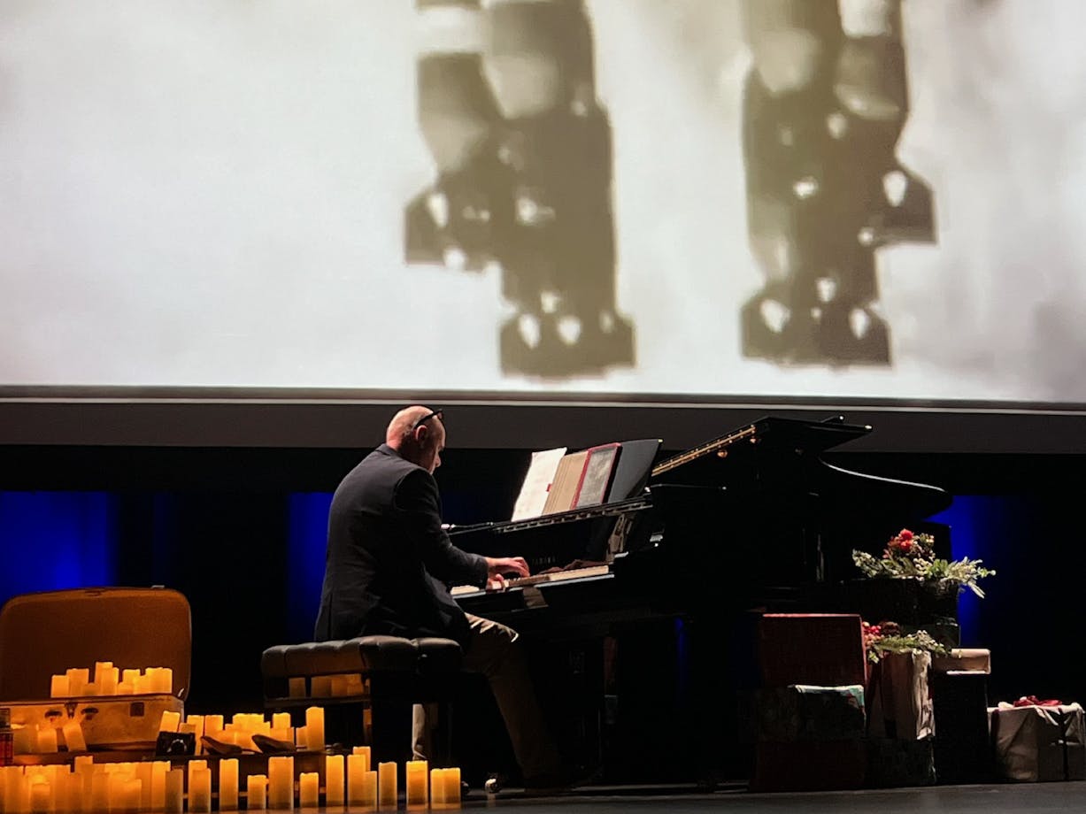Simon Reich playing grand piano in front of screen showing bombs being dropped during World War 2