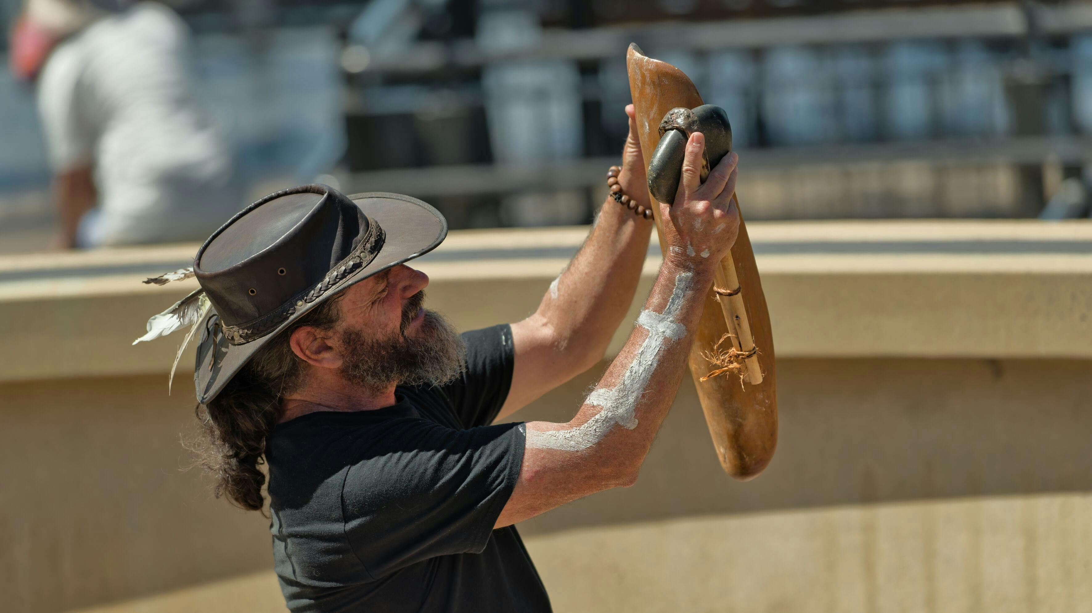 一位头戴帽子、手持传统原住民工具的男子站在风景画前。