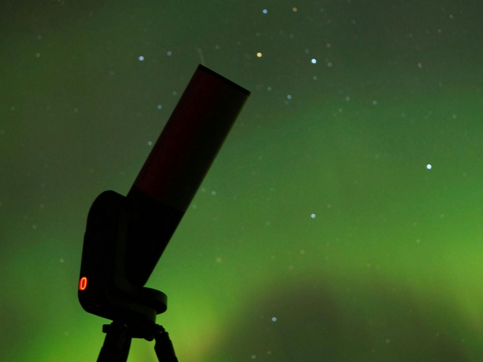 Telescope pointed at the night sky with bright aurora colours glowing above.