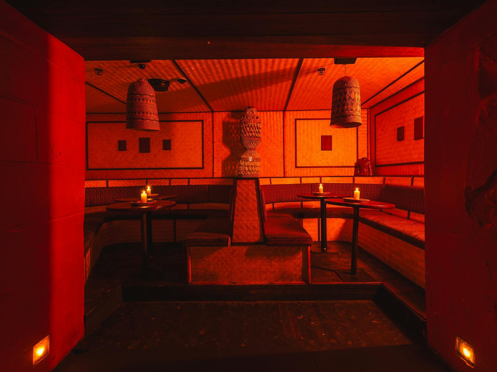 Cozy nightclub booth with bamboo walls, red lighting, bench seating, candles, and woven lamps
