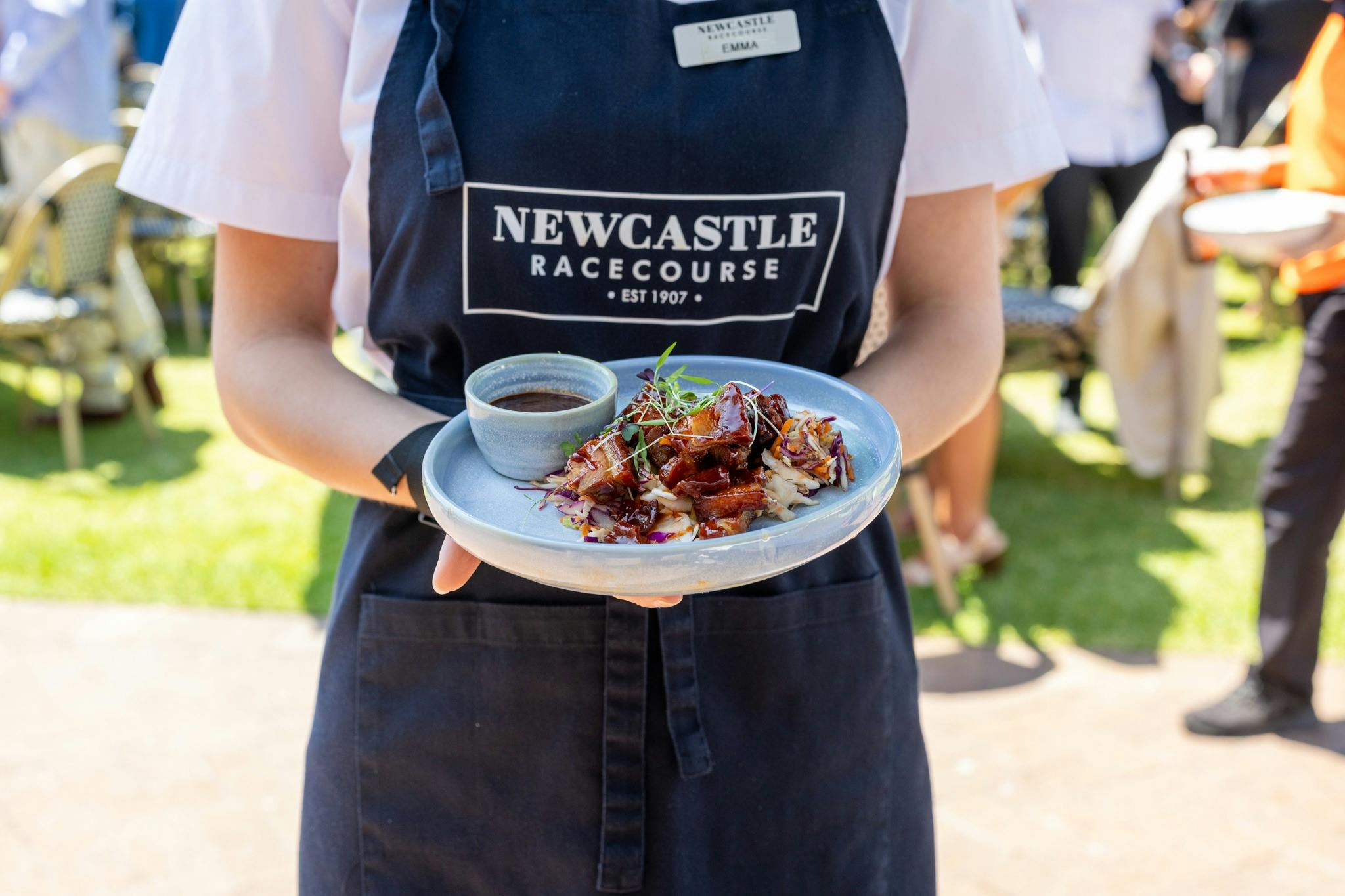 A staff member holding a plated dish outdoors at Newcastle Racecourse.