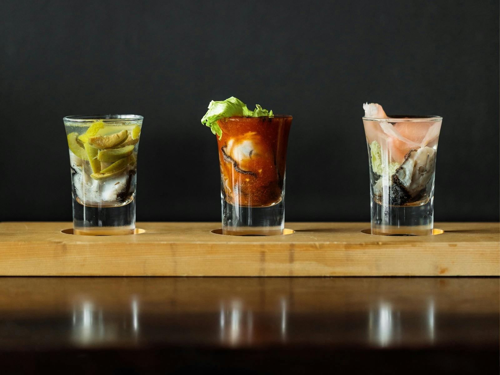 Three shot glasses lined up, each containing an oyster and a different alcohol or garnish
