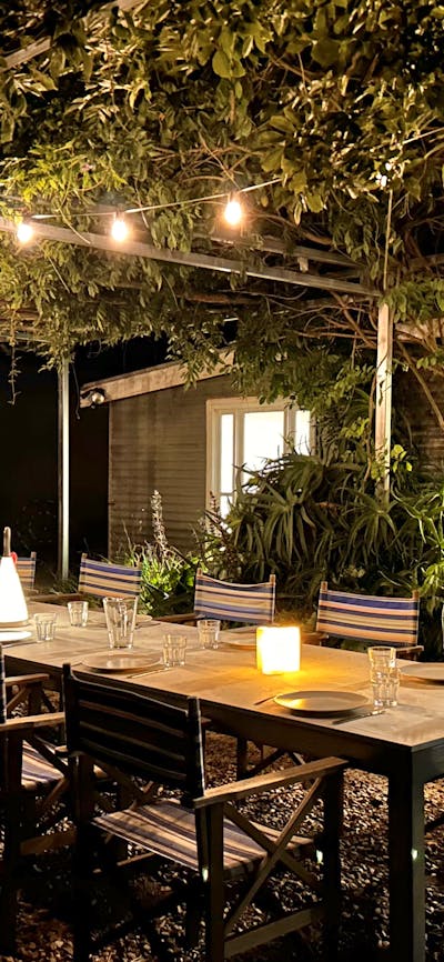 Very long outdoor dining table with candles, lanterns at night. string of lights & vines overhead