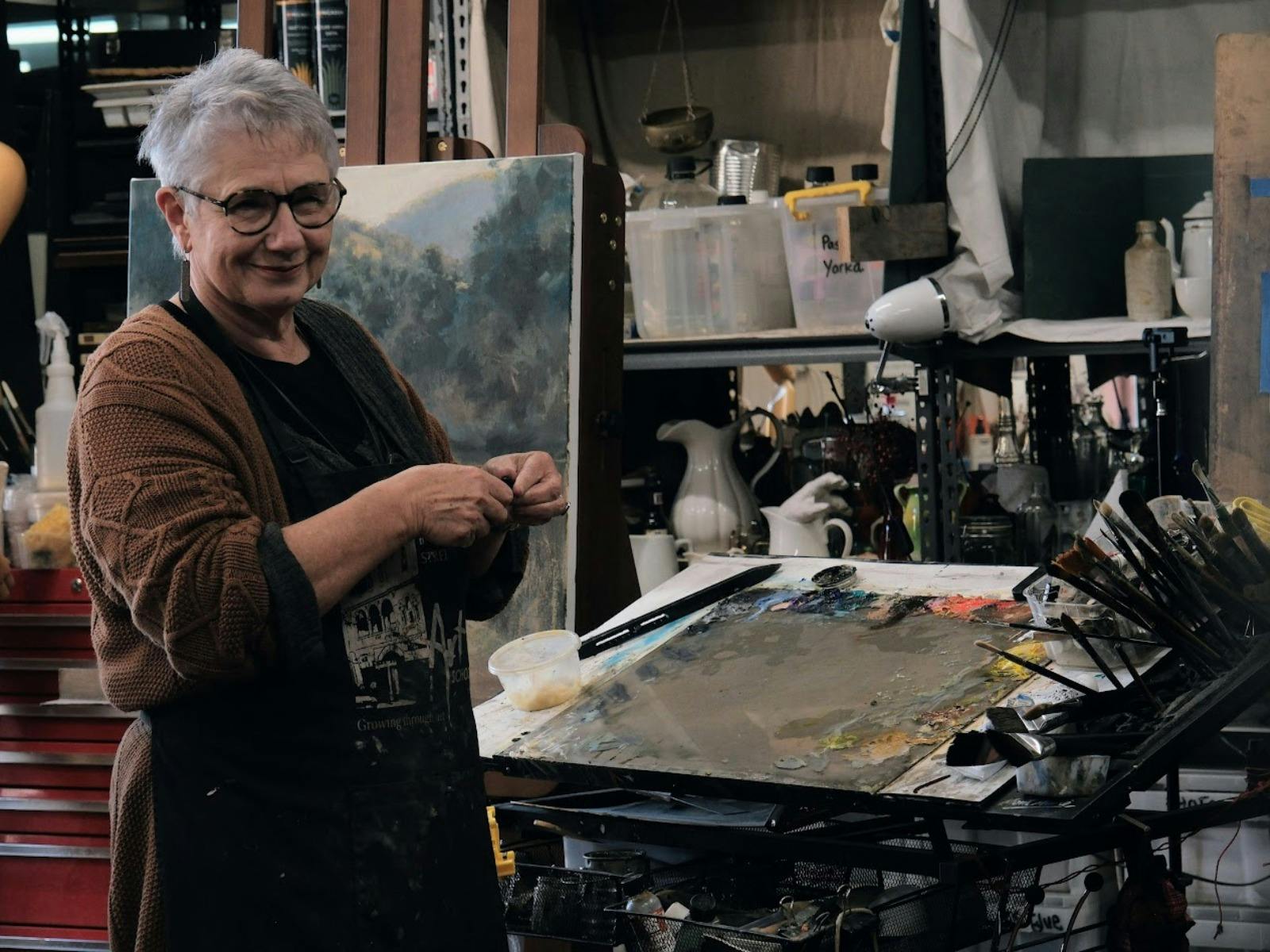 An artist stands at a workbench in her studio, surrounded by brushes, paints and a large canvas.