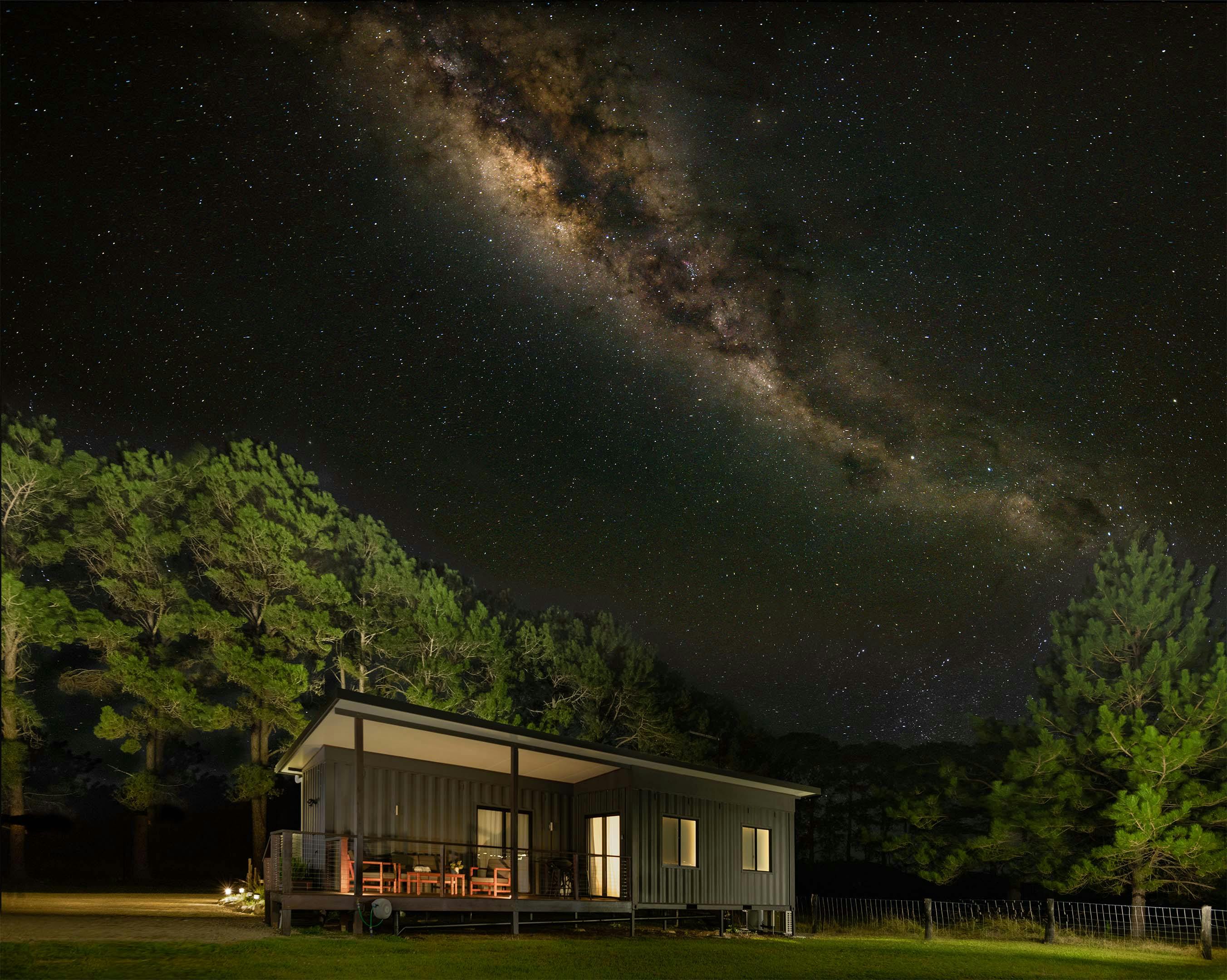 An image of Nightcap cottage sitting in a paddock at night with the Milky Way arching over the top.