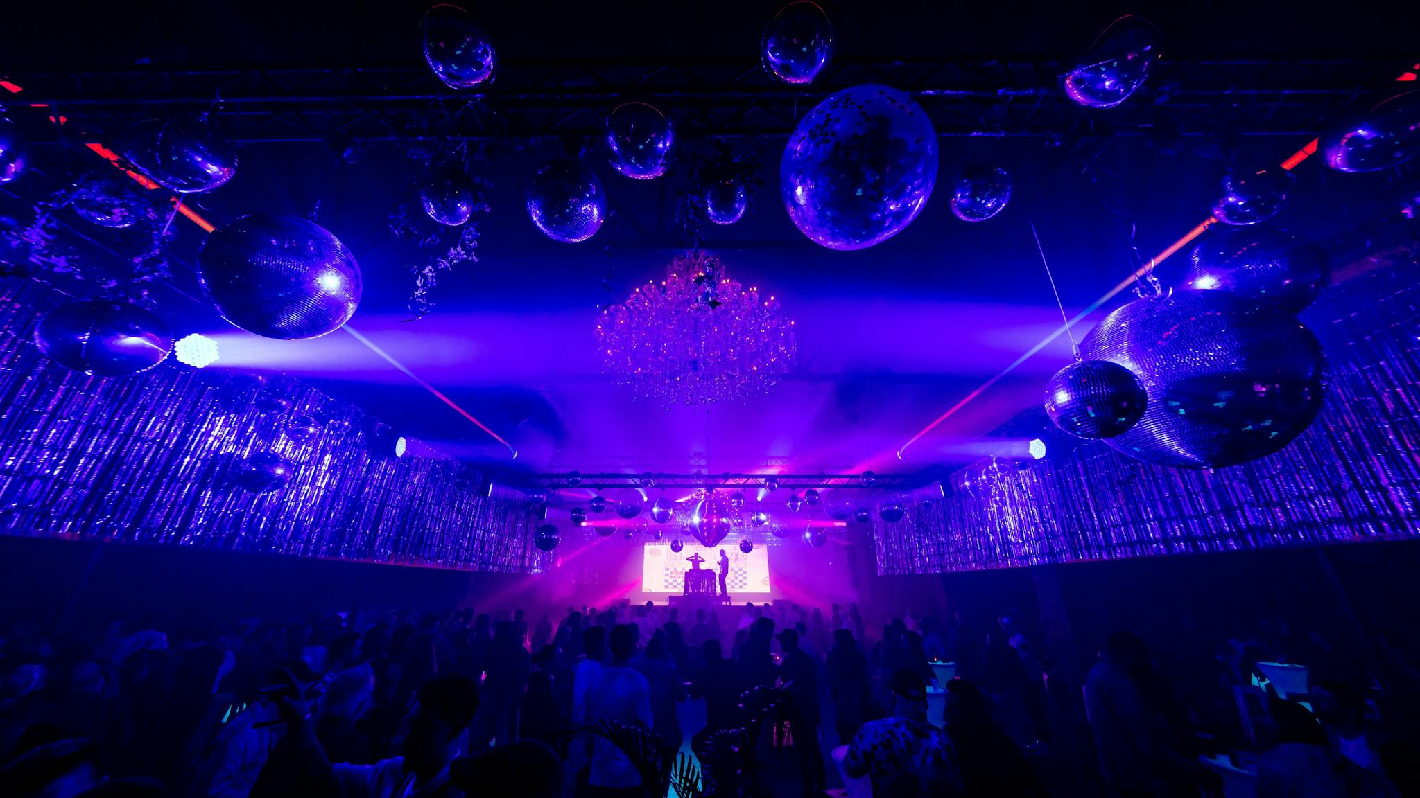 Large mirror balls hang above a crowd as a DJ plays on stage to a crowded room