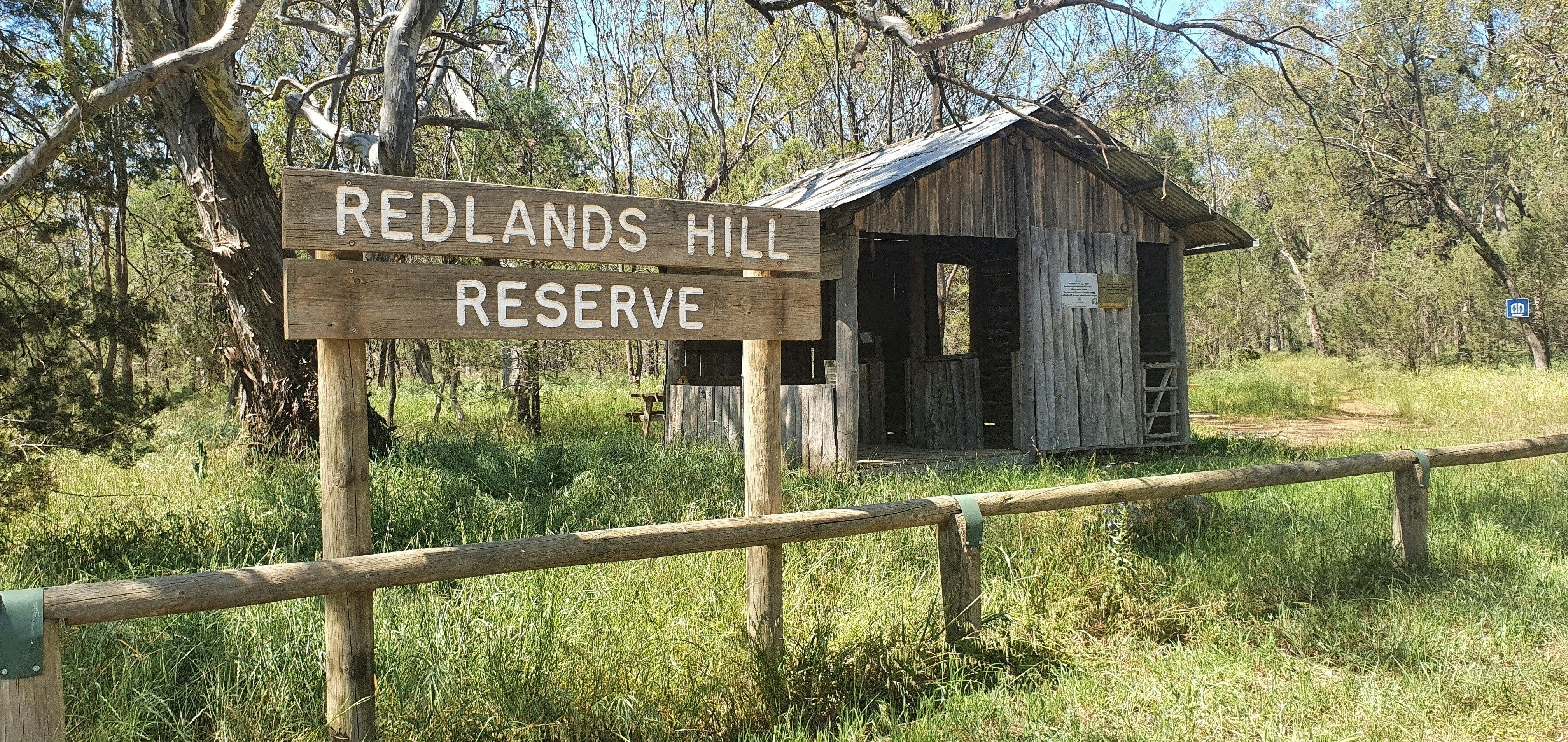 A photo of a wooden sign reading Redlands Hill Reserve. A shearing shed is behind it.